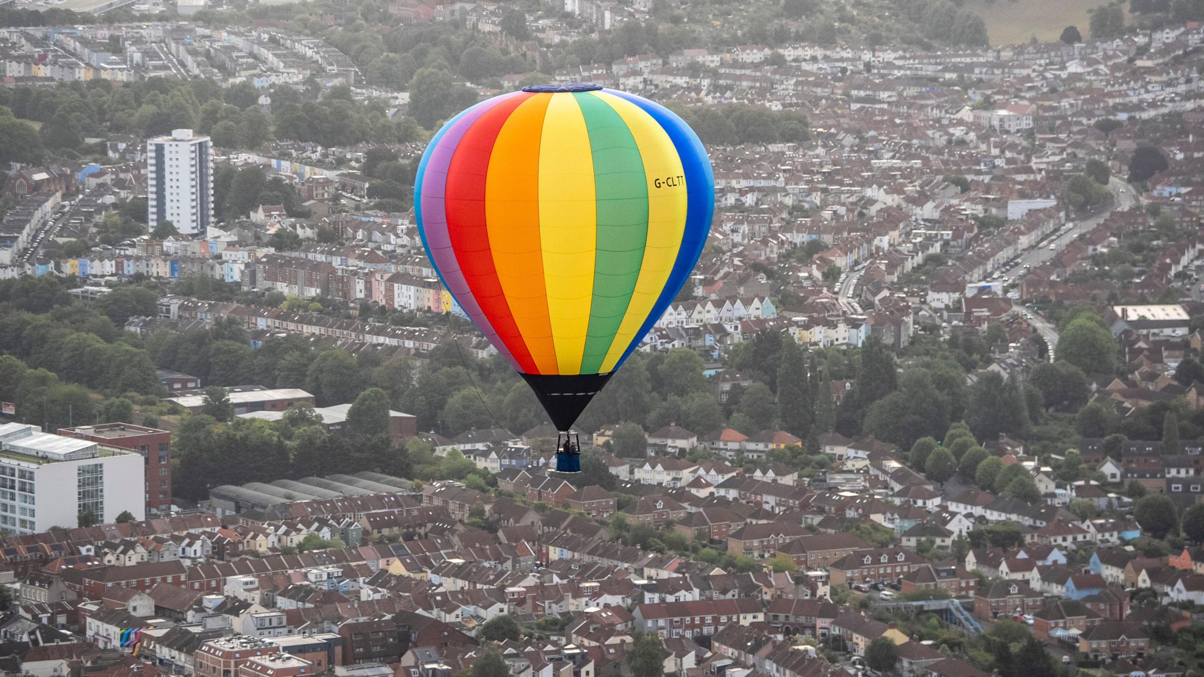 A multi-coloured hot air balloon, with yellow, red, orange, blue, green and purple panels on its canopy, flies over south Bristol. Many rows of terraced houses are visible below it, along with one tower block.