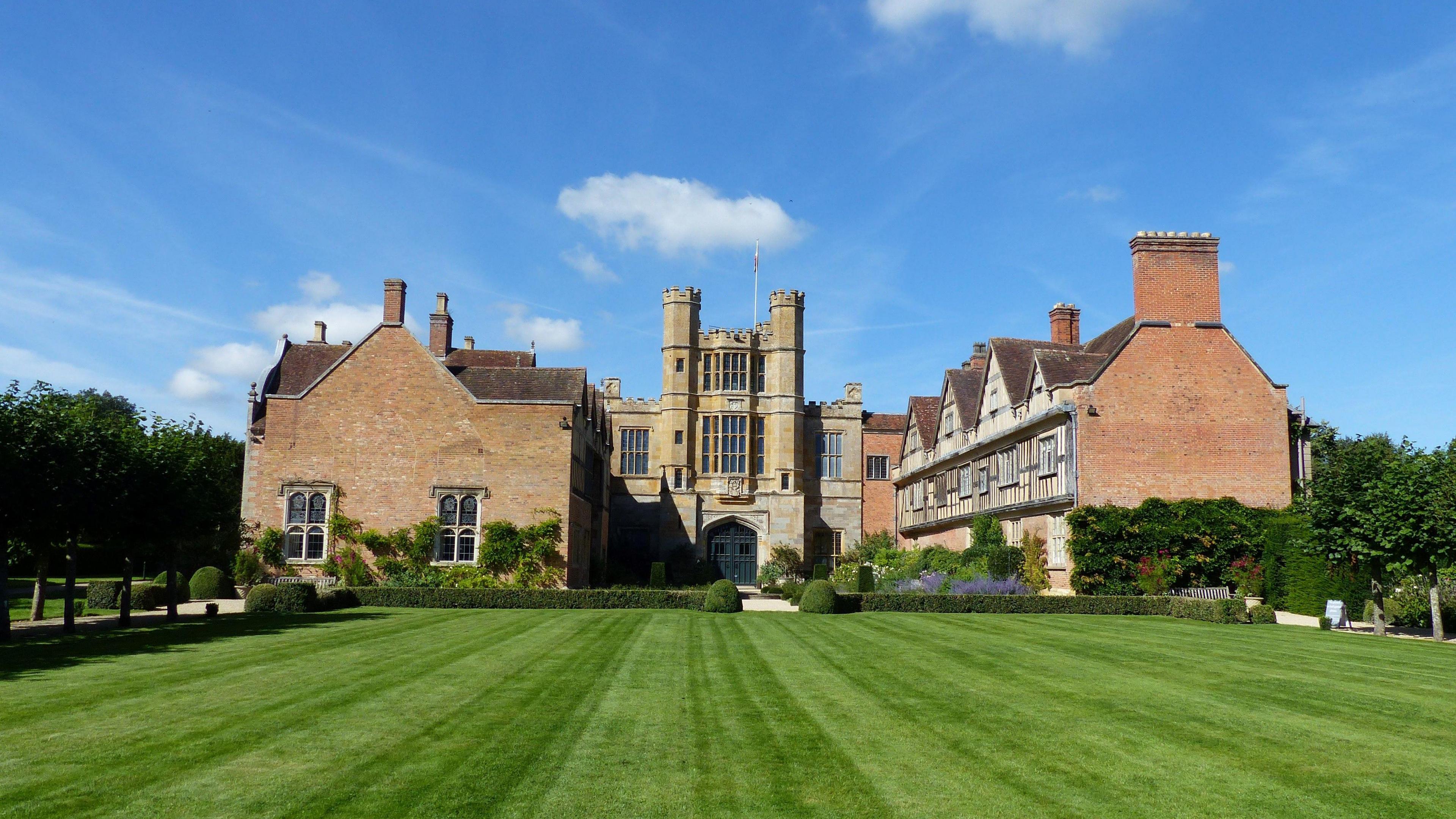 A huge Tudor-style country house behind vast grounds lined with hedges, trees, and with a footpath around the perimeter. 