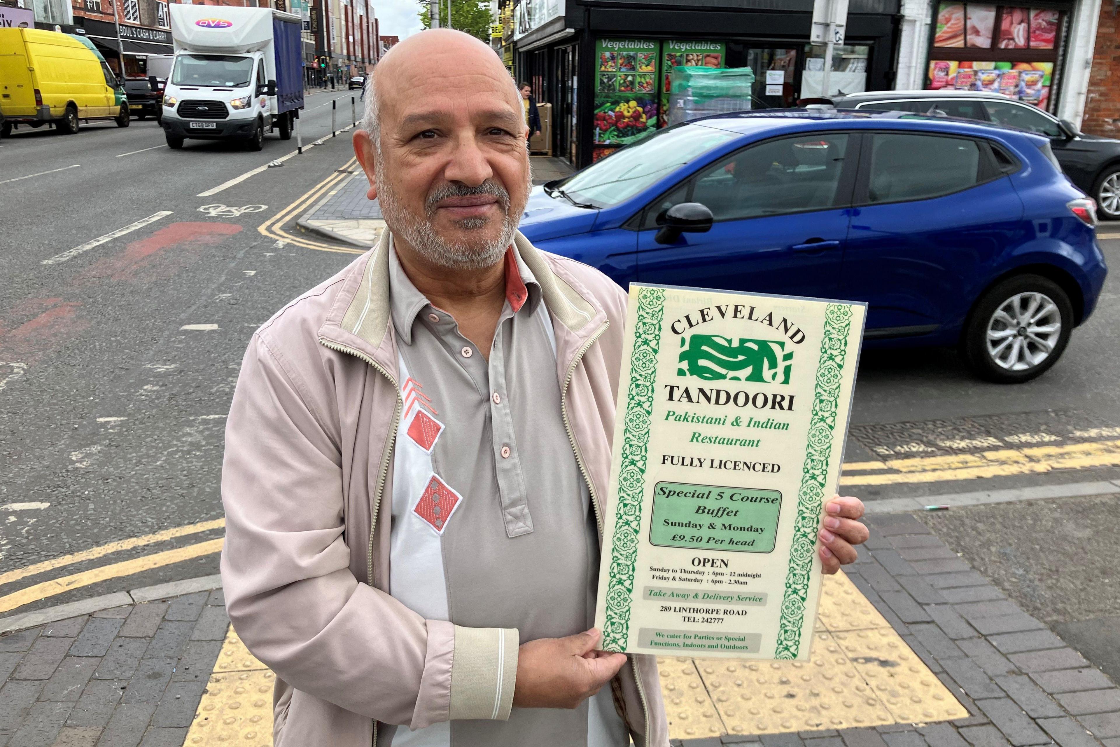 Khadim Hussain standing on Linthorpe Road holding a sign for his old restaurant, Cleveland Tandoori. He is bald with a grey beard and is wearing a grey jacket and polo T-shirt. Vehicles can be seen behind him along with shops.