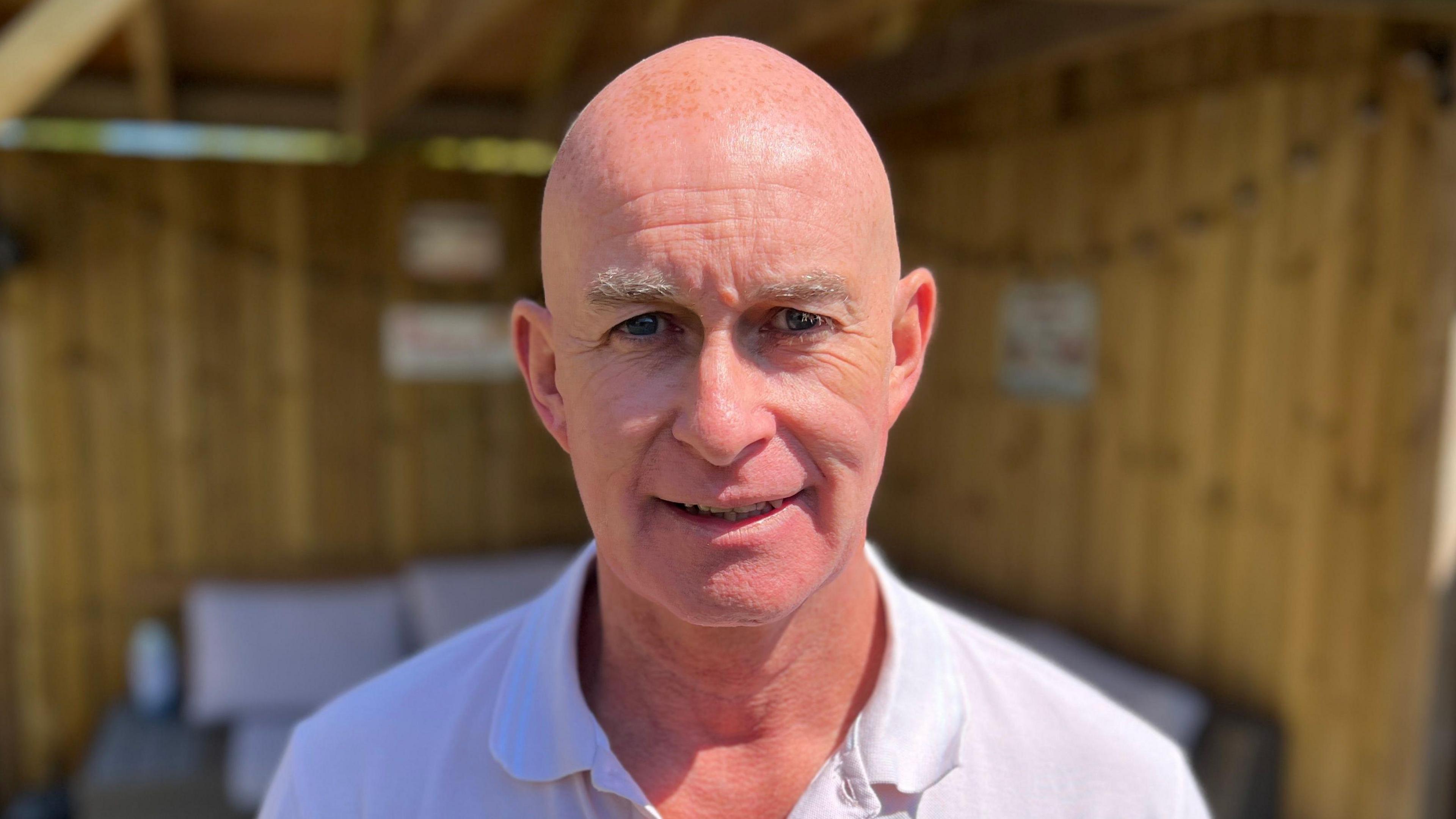 Eamonn Aston smiles into the camera. He's wearing a white polo shirt. He is bald. Behind him is a blurred shed with a visible seating area. 