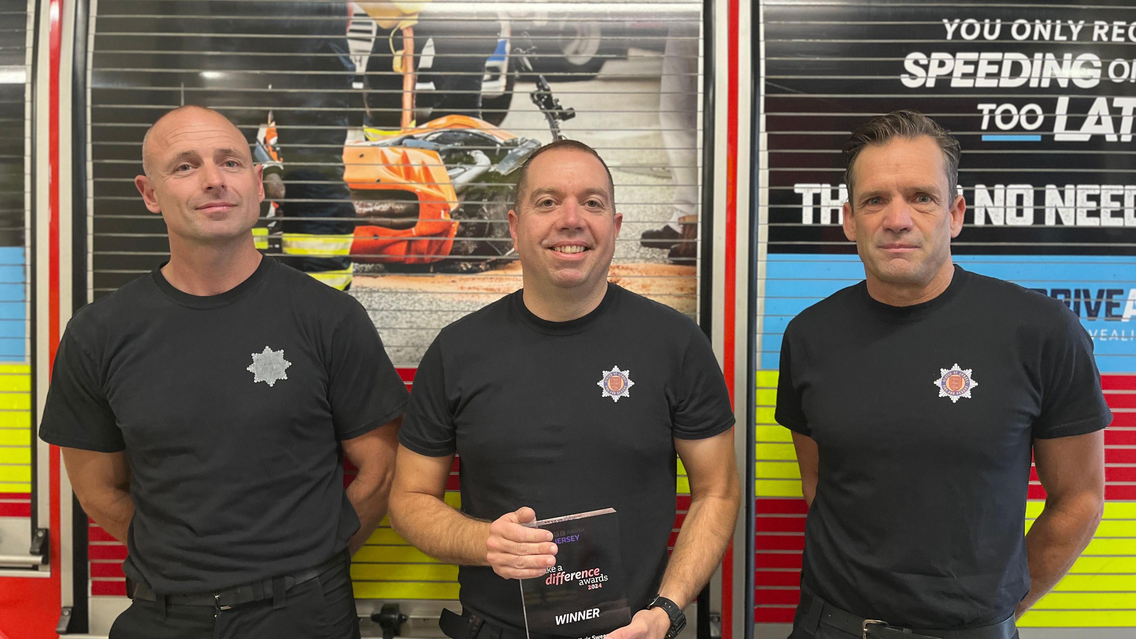 Chris Sweeny (left), Jonny Burch (centre) and Nick Willis (right) all stand in front of a fire engine and look at the camera. They're wearing black T-shirts with the Jersey Fire and Rescue logo on their chests. 