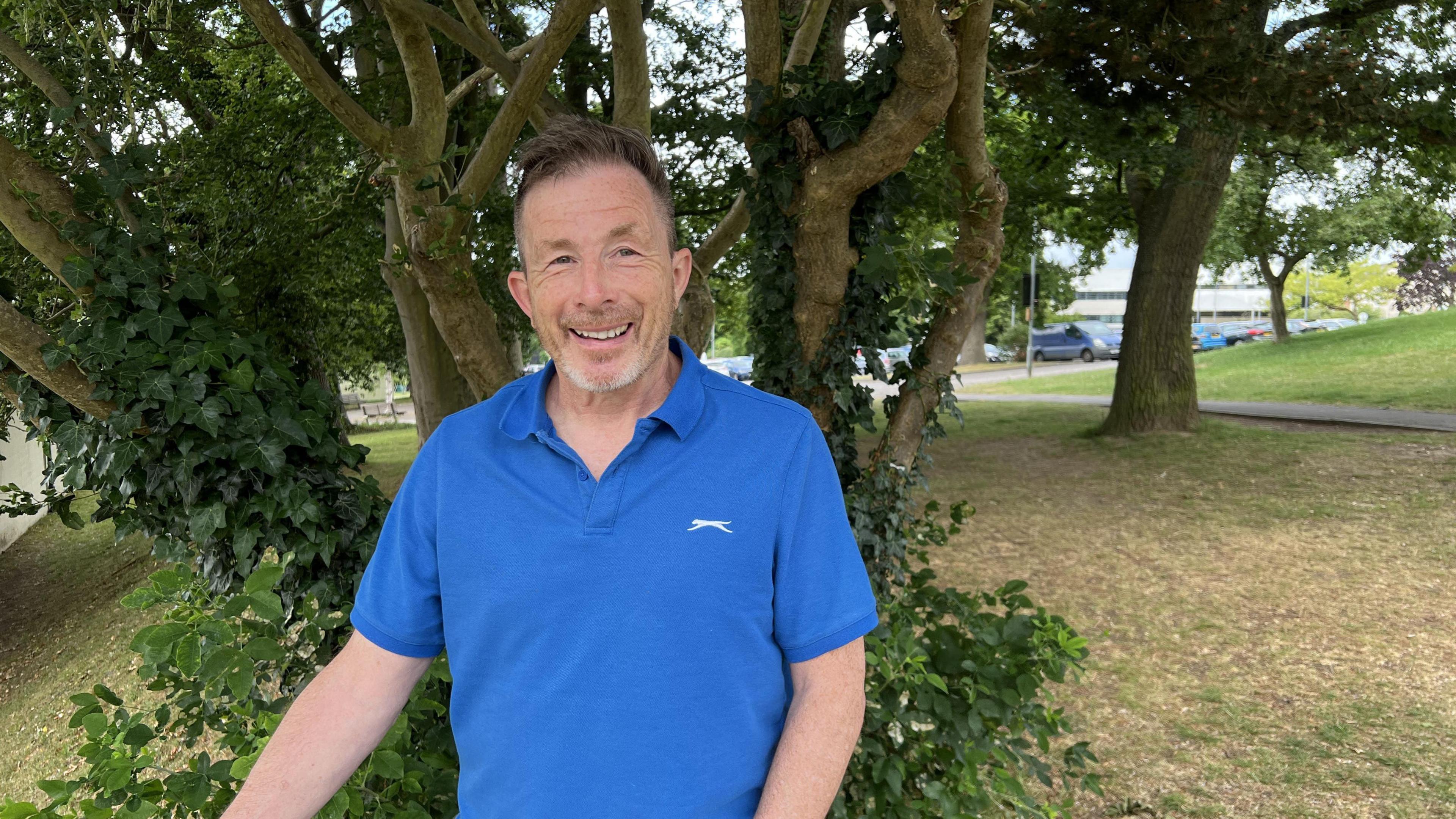 Colin Weston, smiling, in a blue polo shirt in front of a tree covered in ivy in Stratford Park in Stroud. The car park and pedestrian path are visible in the background.