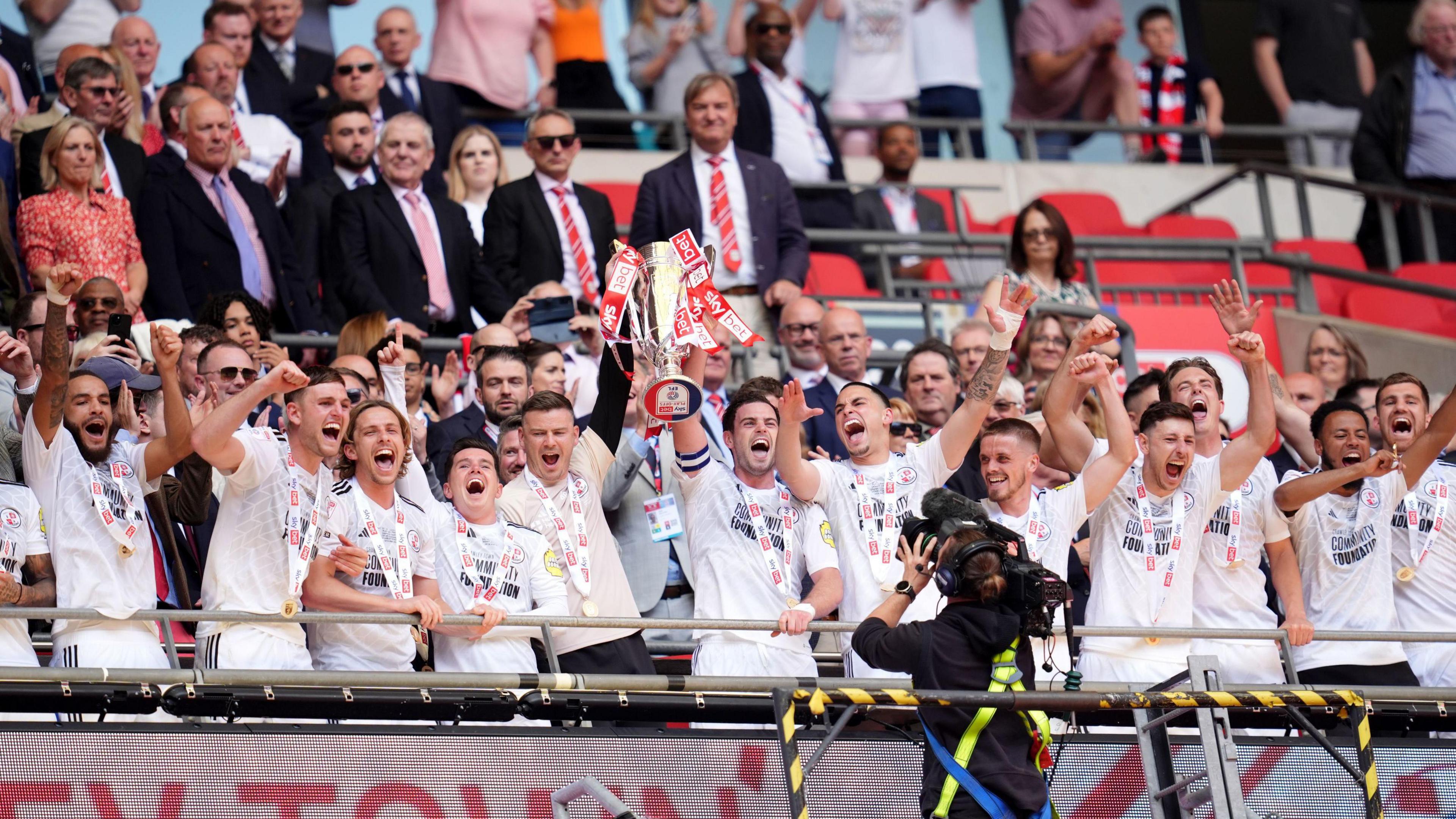 Crawley Town's Dion Conroy lifts the trophy after the Sky Bet League Two play-off final at Wembley Stadium in London. All the players are wearing white tops and medals. 