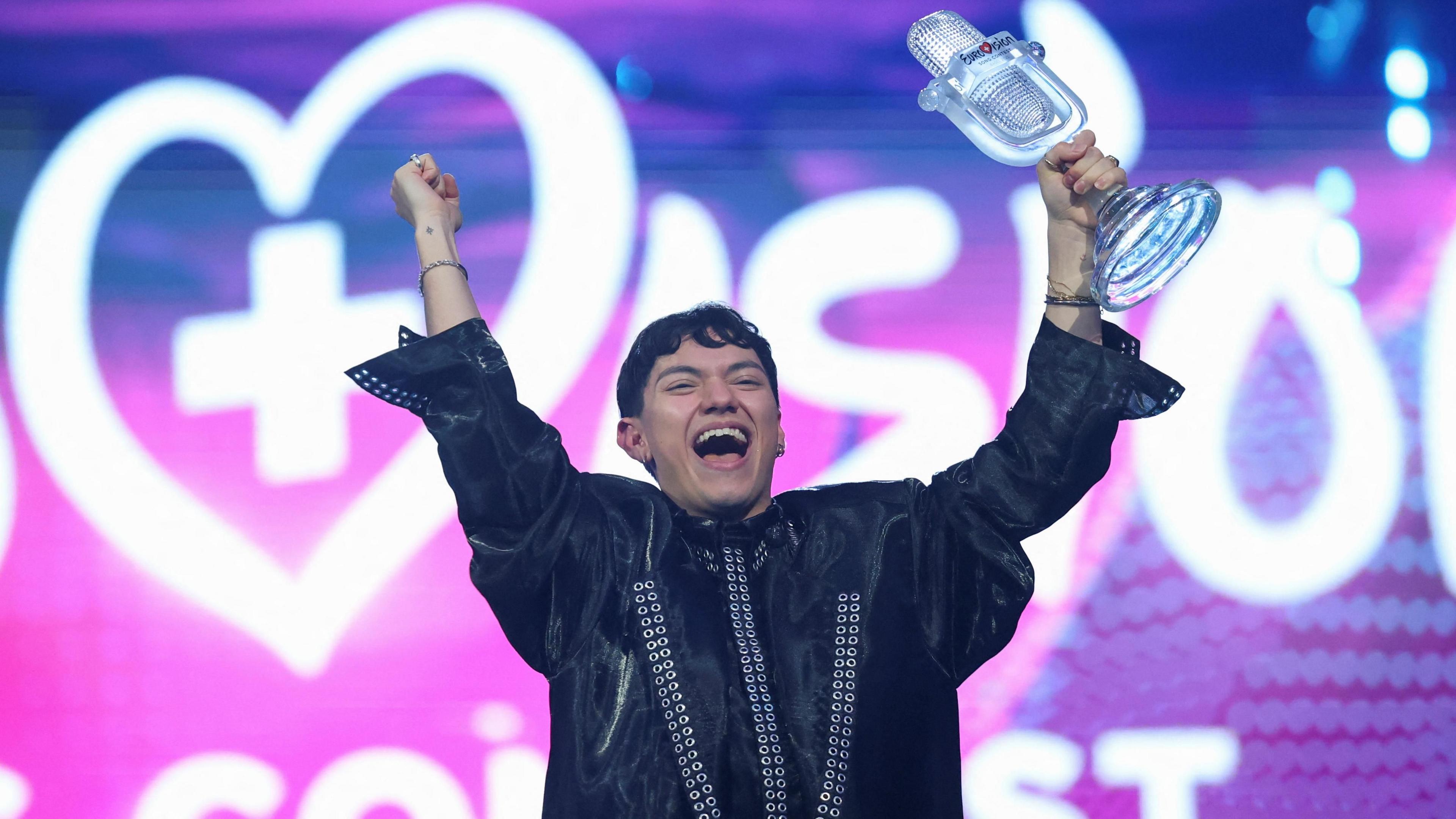 JJ holding a glass microphone trophy aloft in celebration with his other hand in the air against a purple and pink background 