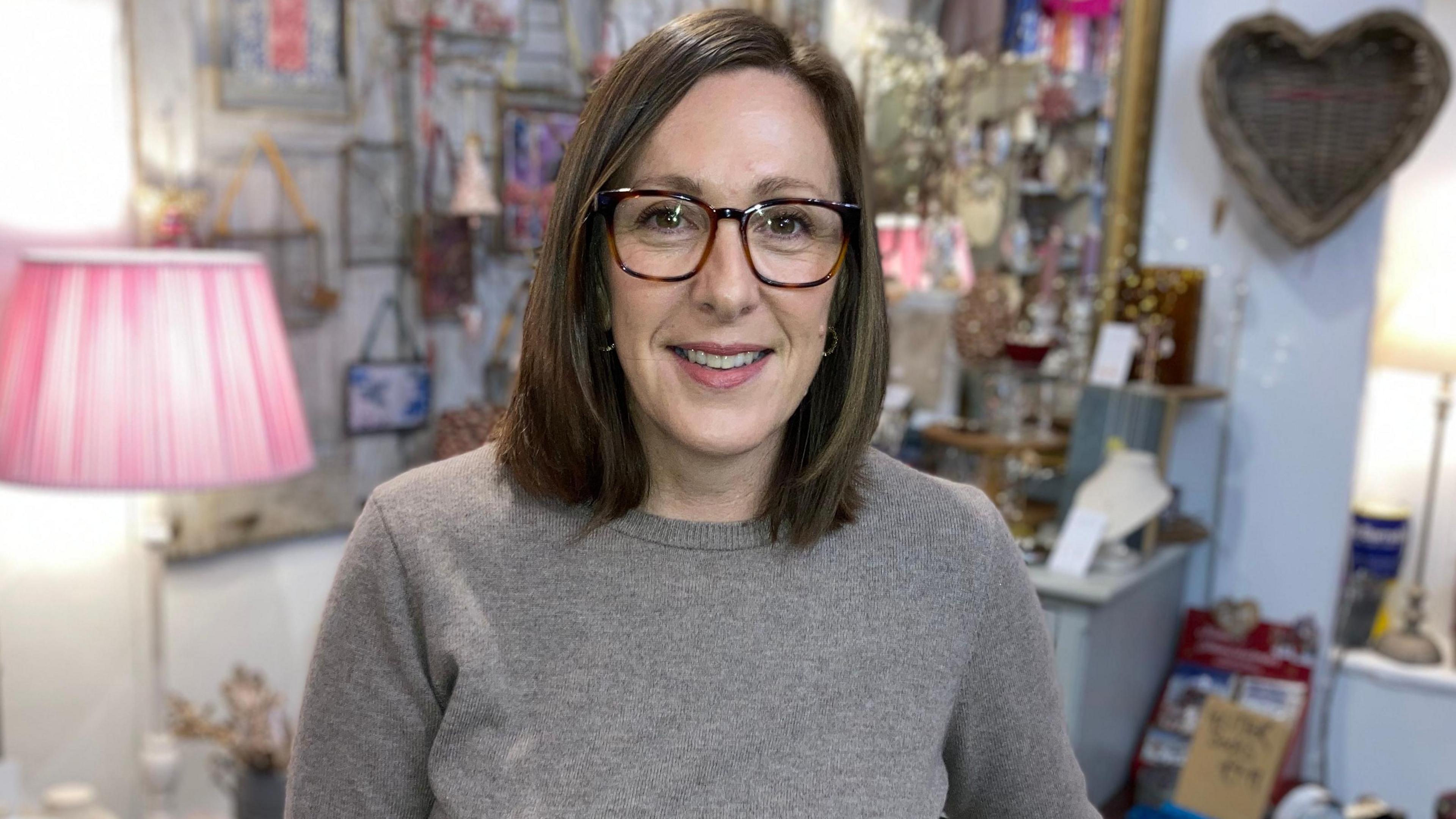 A smiling Josie Rossington wearing a grey jumper and glasses, standing in front of a display in her gift shop in Lincoln