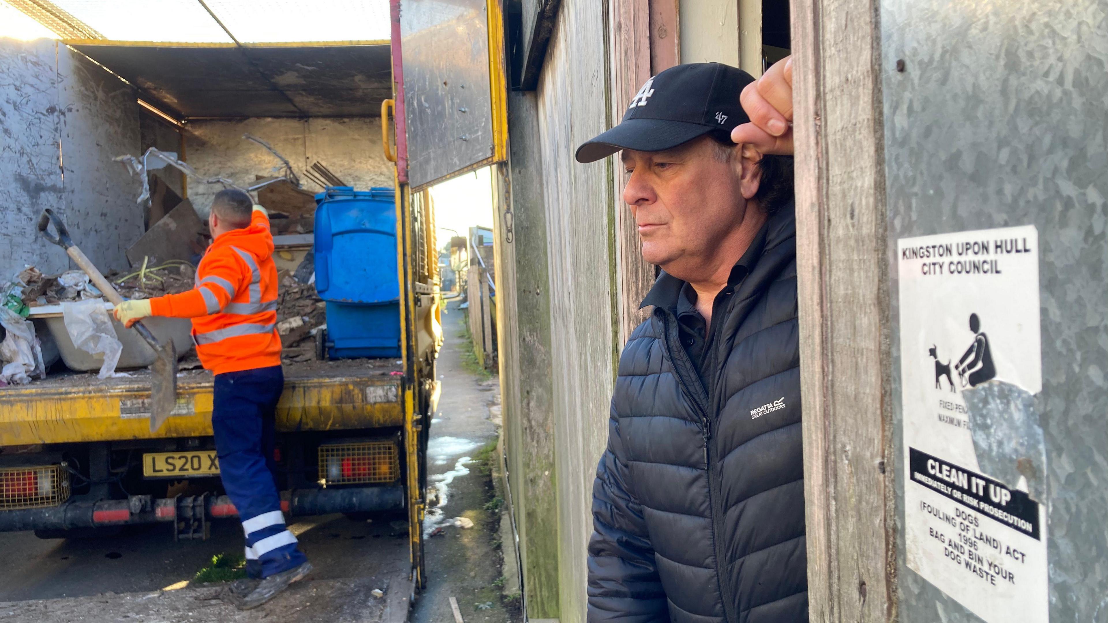 A casual side profile of Darren Speck, wearing a black cap and a black padded Regatta coat. He is leaning against a door frame with his left hand. In the background is a man in blue trousers, black boots and an orange hi-vis jacket putting the rubbish into a yellow van. 