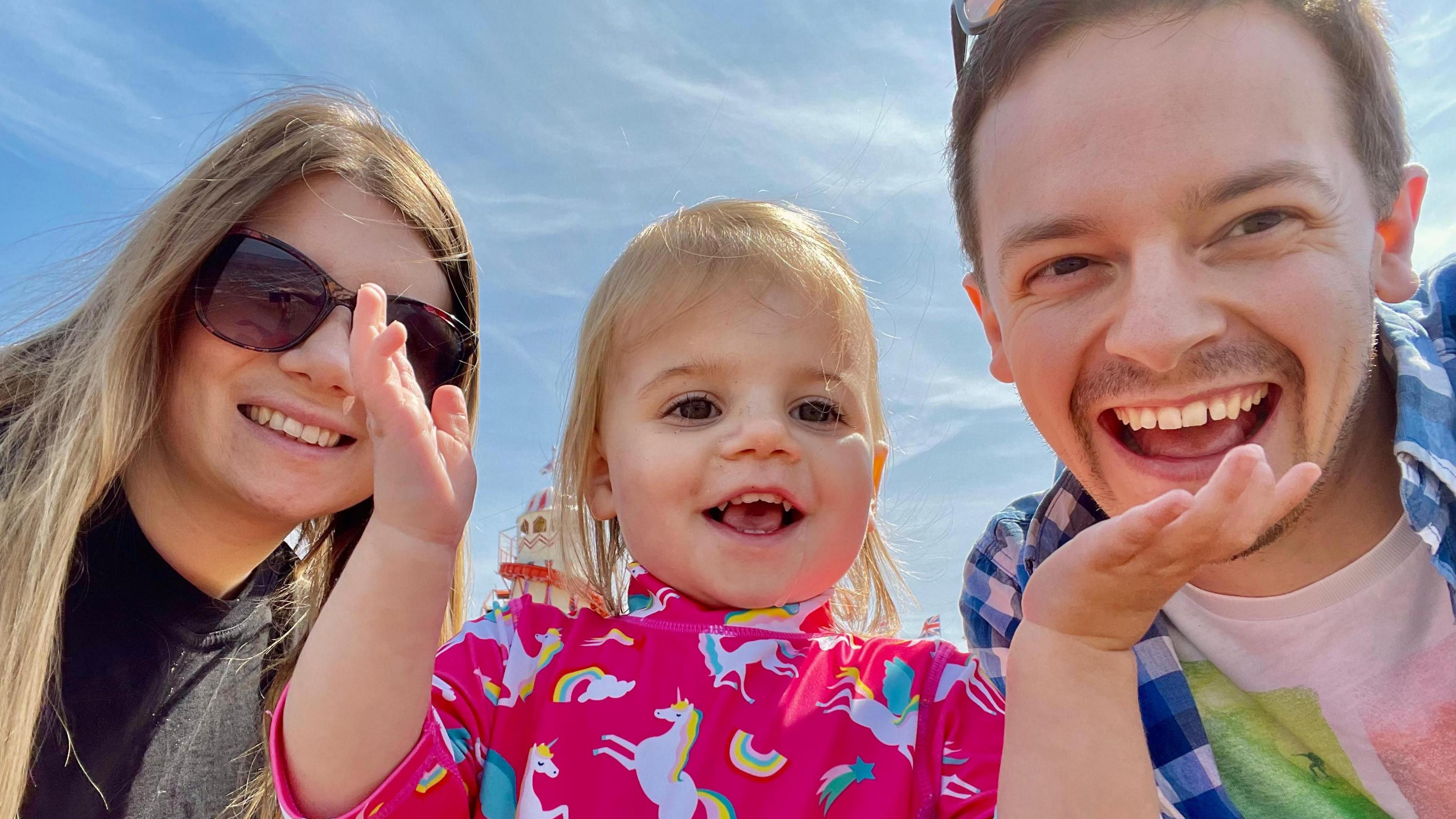 A woman and a man posing for a photo with a smiling, female toddler between them. The toddler has her hands raised in the air. The woman is wearing sunglasses. They are set against a bright blue sky.