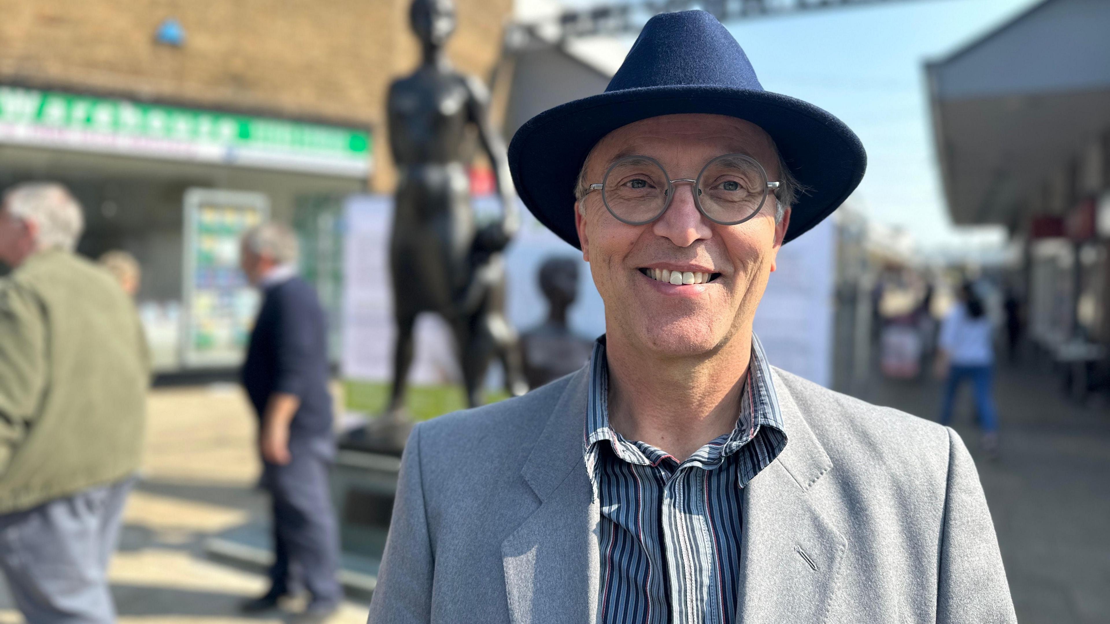 A head and shoulders shot of Ceri Thomas. He is wearing a grey jacket, circular glasses and a navy fedora style hat