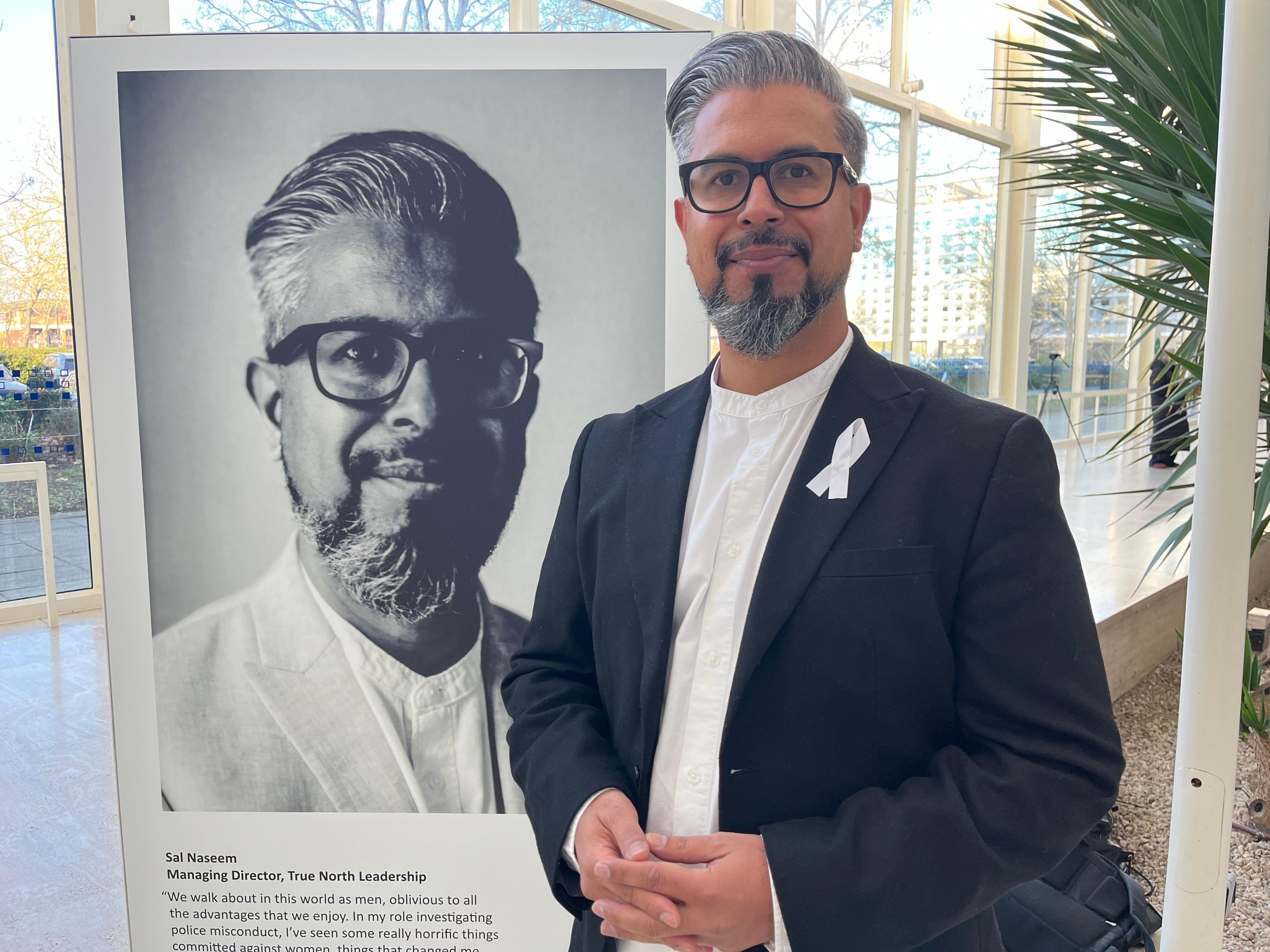 Sal Naseem, who is wearing a dark suit jacket with a white ribbon pinned to the lapel and a white collar-less shirt. He is standing beside the portrait of him that features in the exhibition. He has swept-back grey hair, a grey beard and is wearing glasses. He has his hands clasped in front of him. The picture is taken in a large glass-sided room. The leaves of a large plant can also be seen in the background.