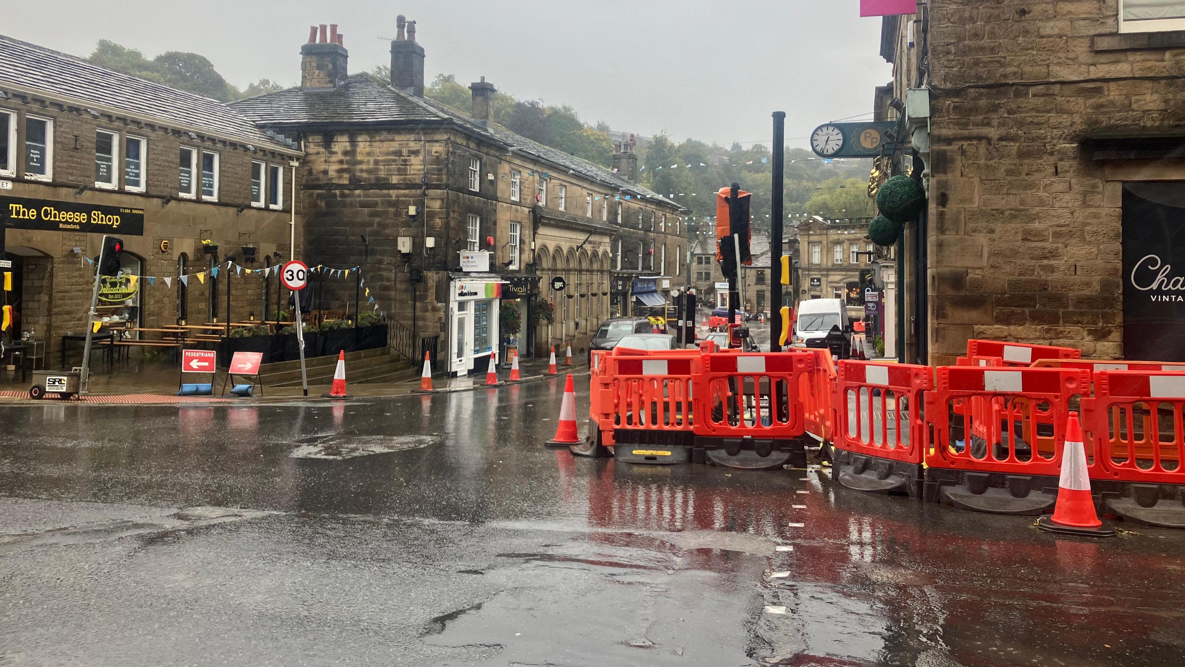 A wet road with bright orange barriers bordering the pavement. Traffic cones line the road and diversion signs are situated alongside.
