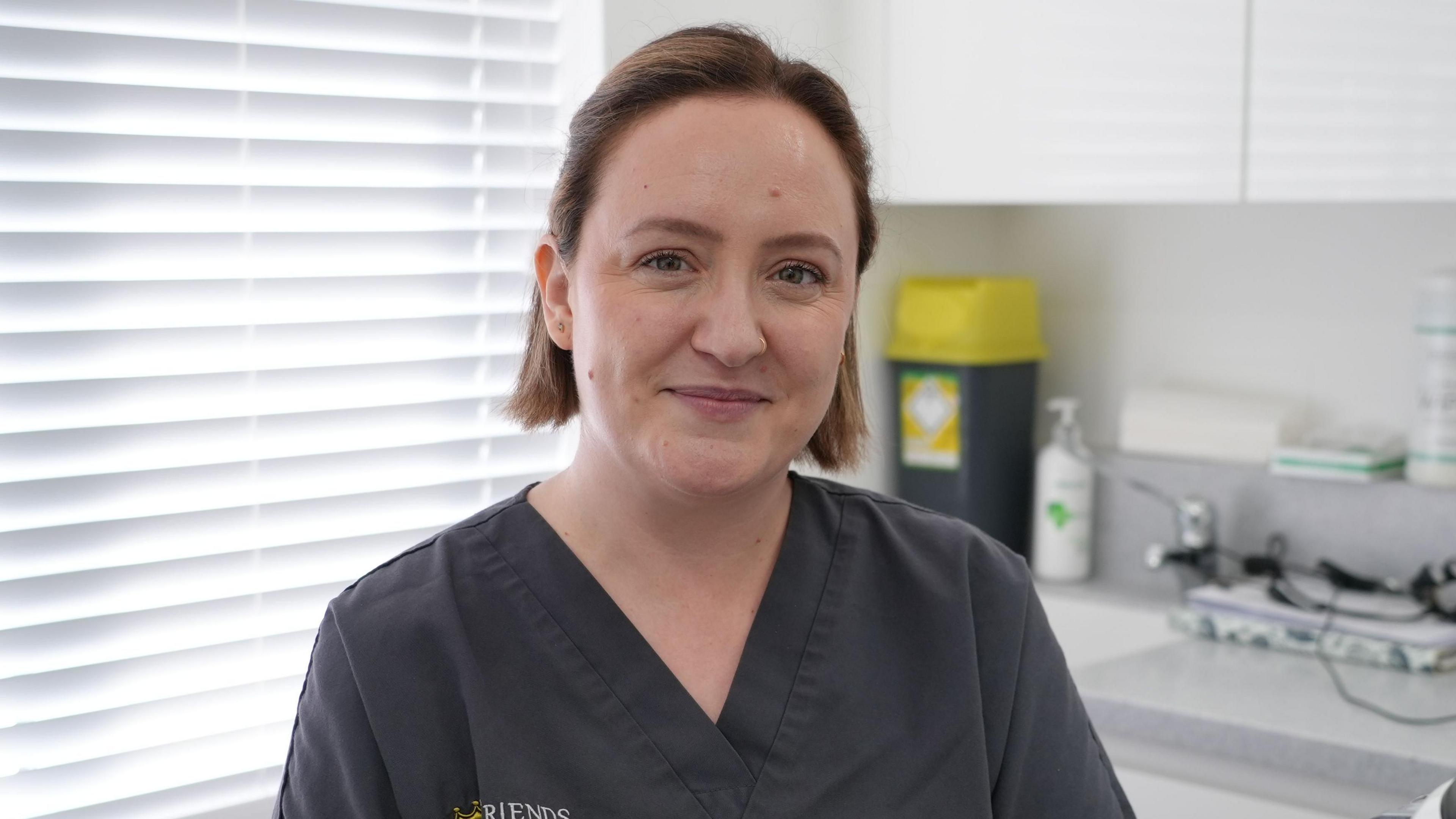 Dr Alba Torrado-Varela, a dentist stood in a dental practice room and is wearing dark grey overalls. She has short brown hair and is looking and smiling towards the camera. 