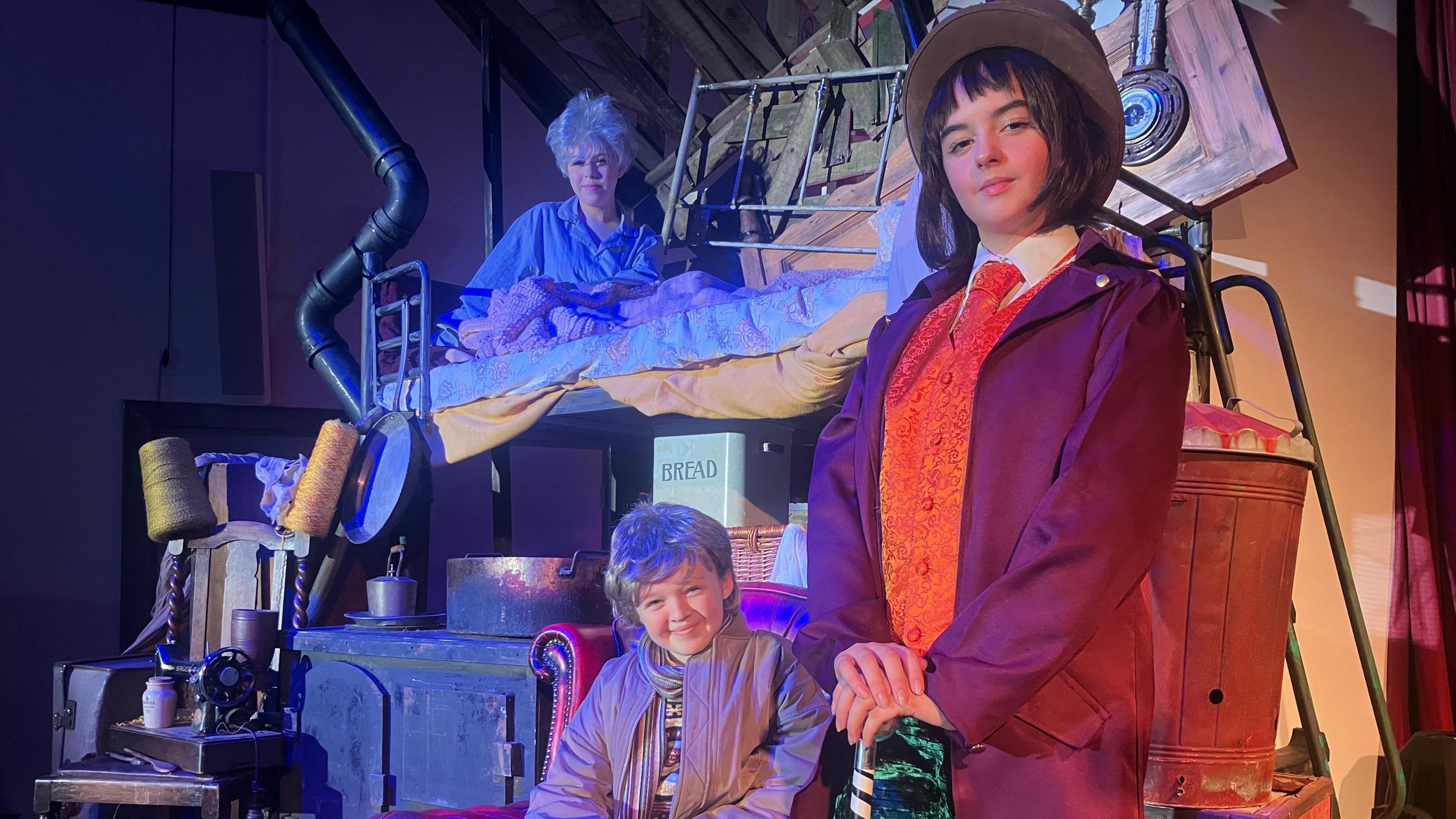 Three children dressed up. One is dressed up as Willy Wonka and is wearing a top hat. One is dressed like Charlie and is wearing a blond wig. The other is lying on a prop of a bed and is wearing a white wig. Various household props are onstage, like a bread container and a chair.