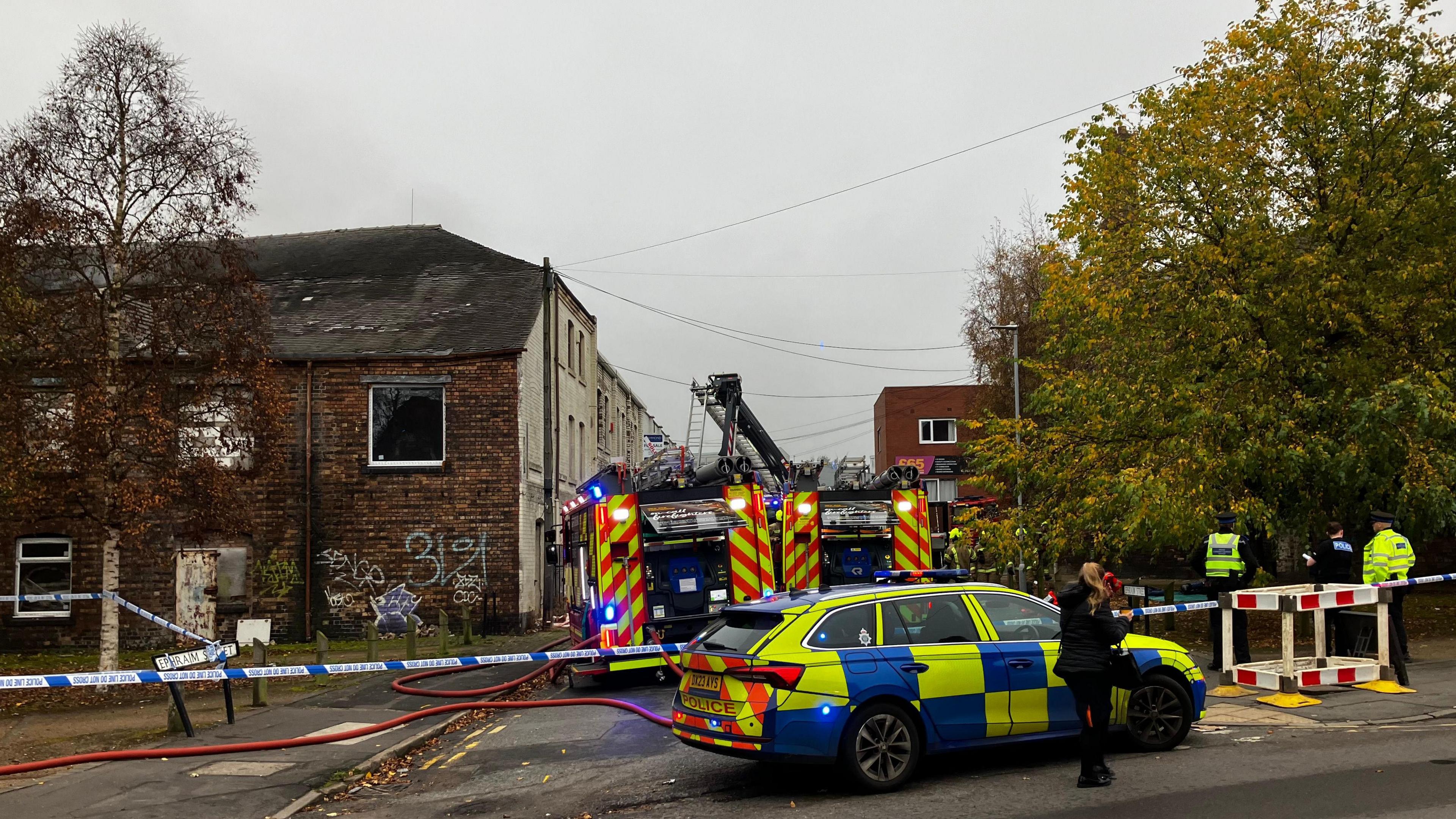 Firefighters and police officers work behind a cordon with police tape blocking off the area where two fire engines are situated. Several police officers can be seen to the right next to a large tree.