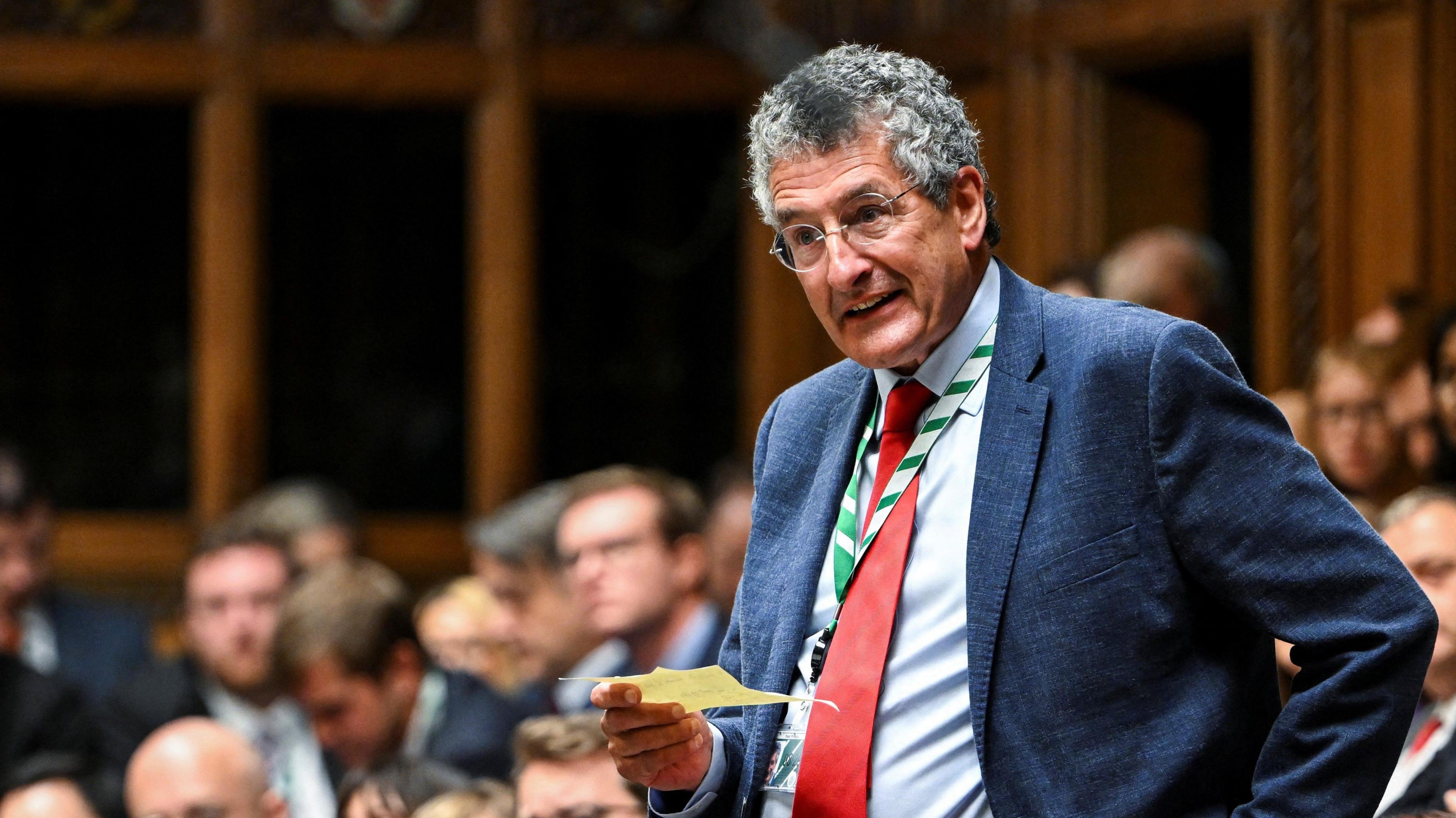 Labour MP Peter Prinsley pictured speaking in the House of Commons during Prime Minister's Questions. He is wearing a blue suit with a lighter blue shirt underneath and a red tie. He is holding a yellow piece of paper.