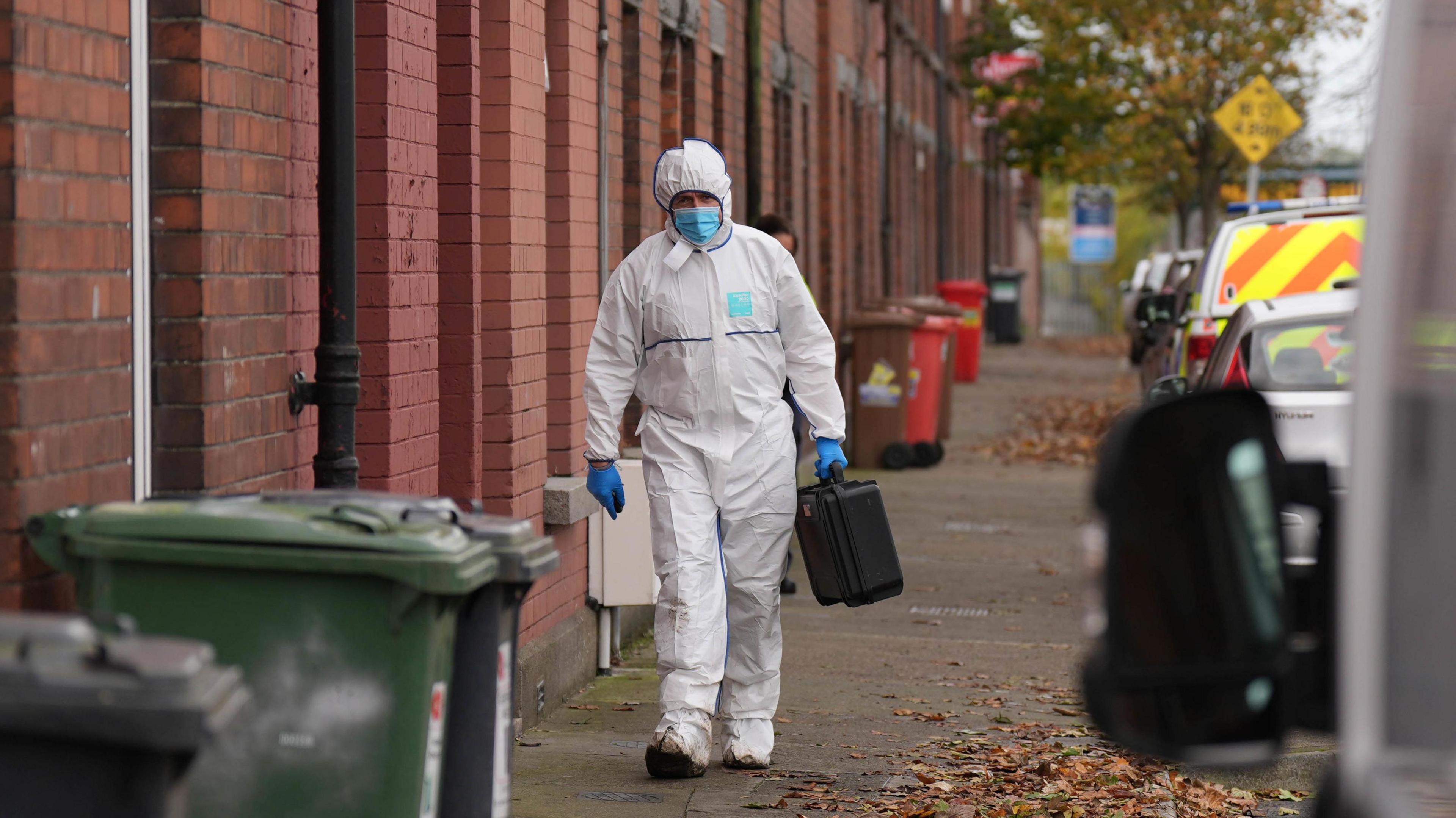 A Gardai forensic officer walking towards the camera on Emer Terrace, a street with red brick terraced houses and lined with parked vehicles. The officer is wearing white hasmat suit and a blue mask and gloves and is carrying a black case. 