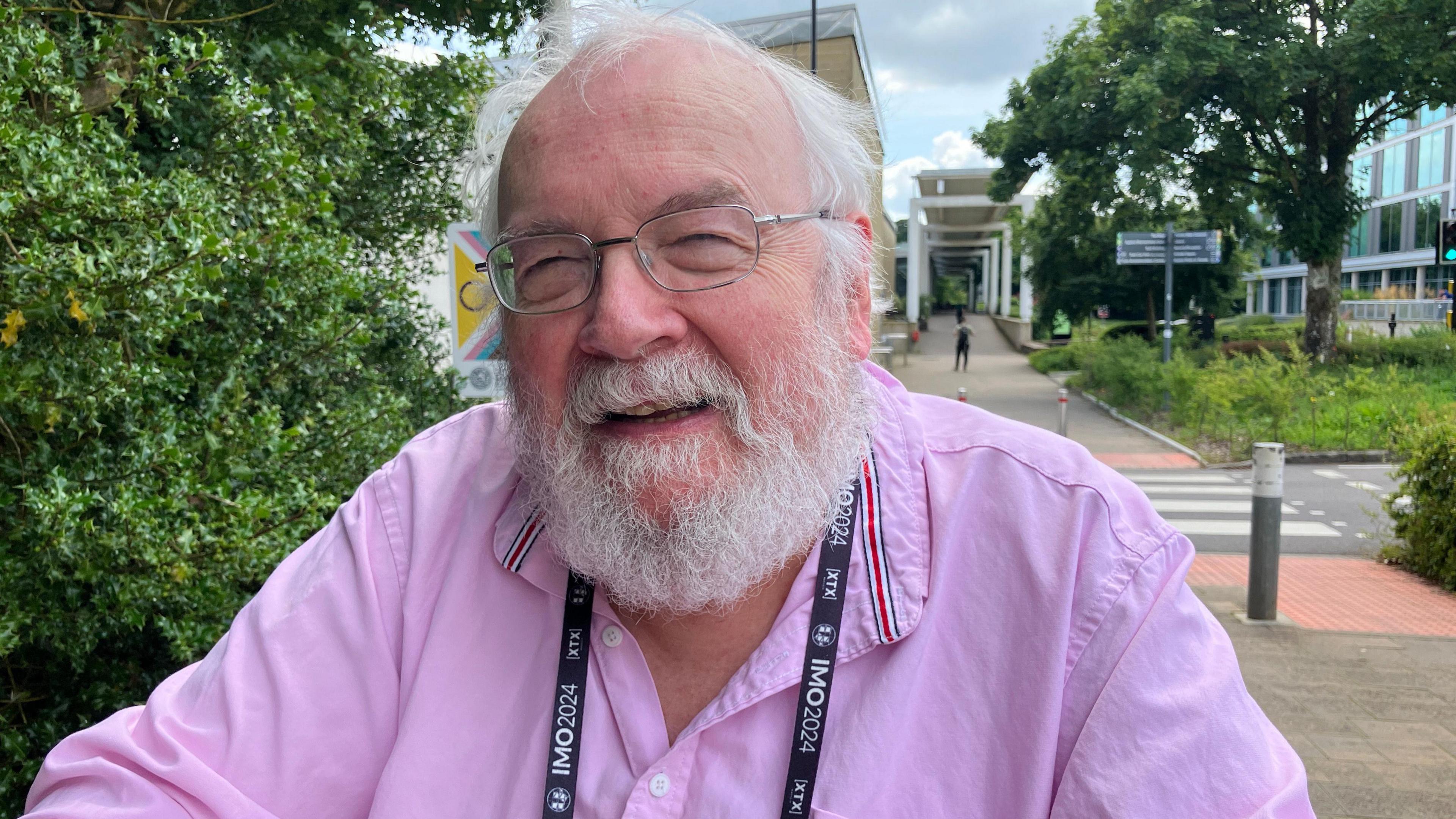 Professor Geoff Smith MBE. He has white short hair and facial hair. He is wearing a pink shirt and glasses. He is looking at the camera and smiling. 