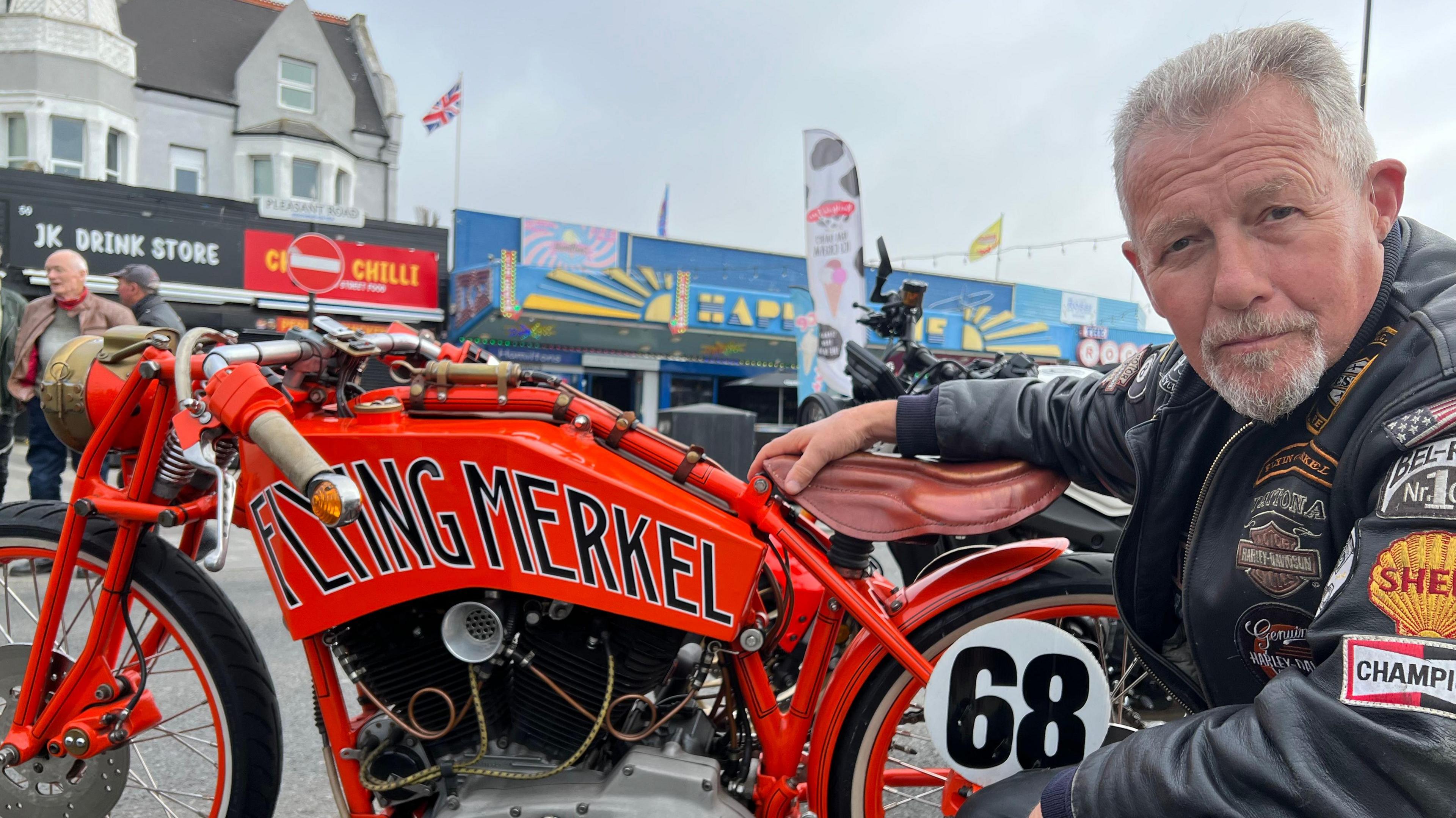 Mark Gelsthorpe kneels to the side of his bright orange bike. He has short grey hair and wears a leather biker jacket with various badges on the arms and chest.