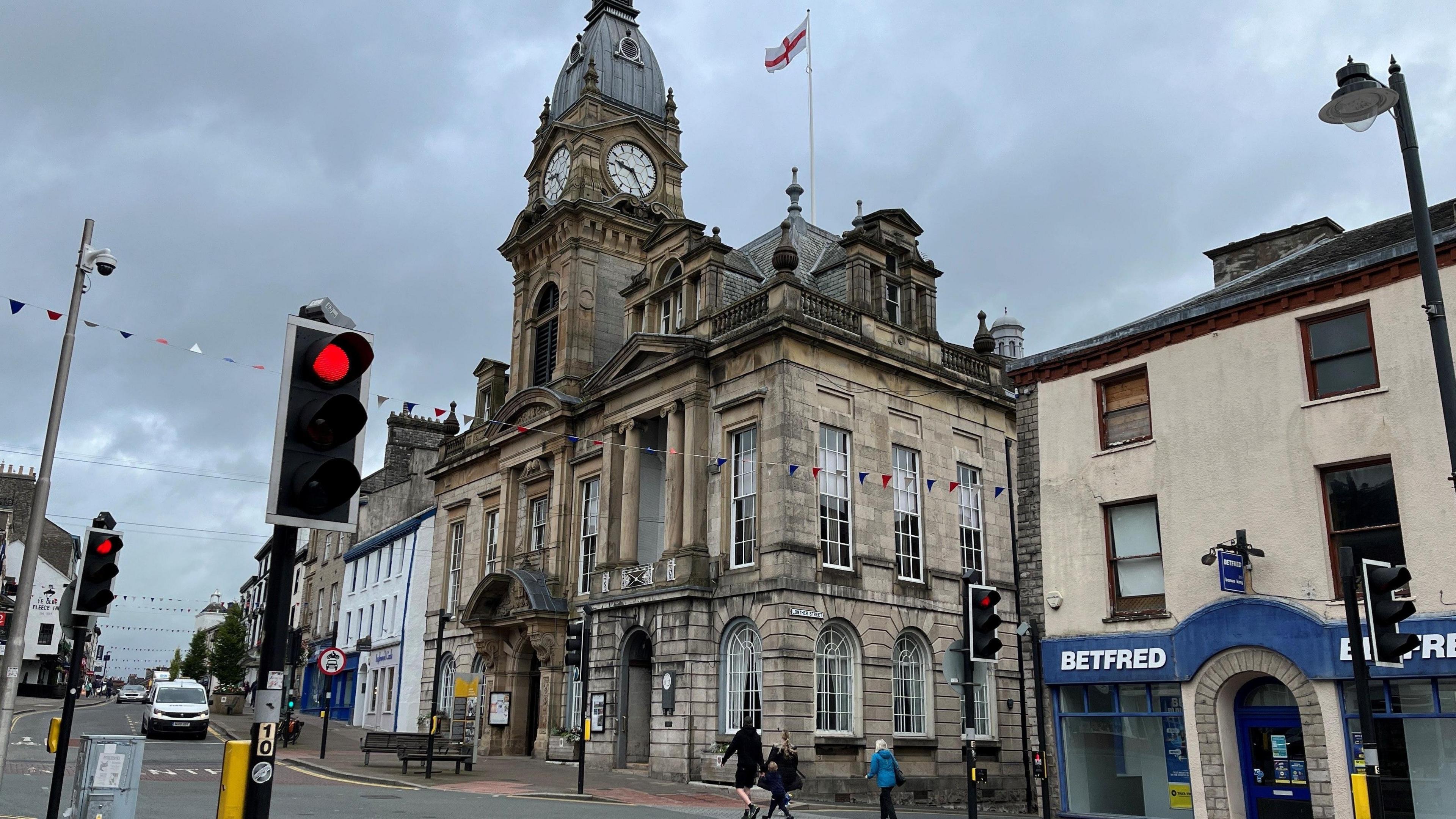 Corner view of Kendal Town Hall, a Gothic revival style building with a clock tower and ornate entrance. A Betfred establishment is to its left and to the right further shops can be seen lining the roadside. Traffic lights - on red - are on the adjoining junction.