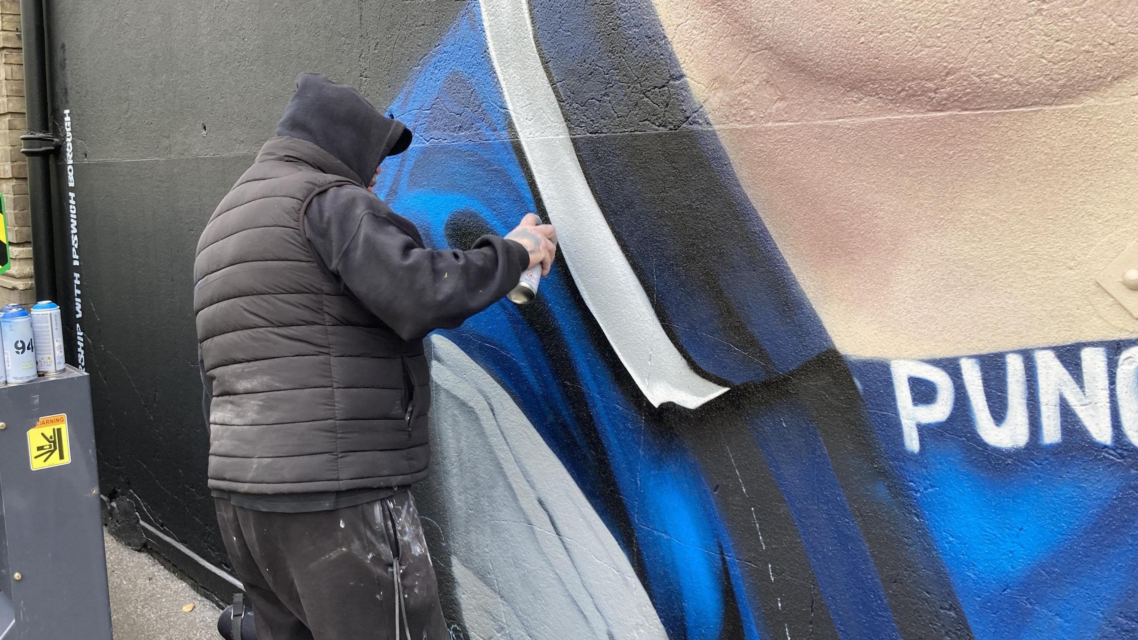 A gentleman wearing a black hoodie, black cap and black puffer jacket is pictured spray-painting the mural. He is painting the collar of Sheeran's football top on the mural.