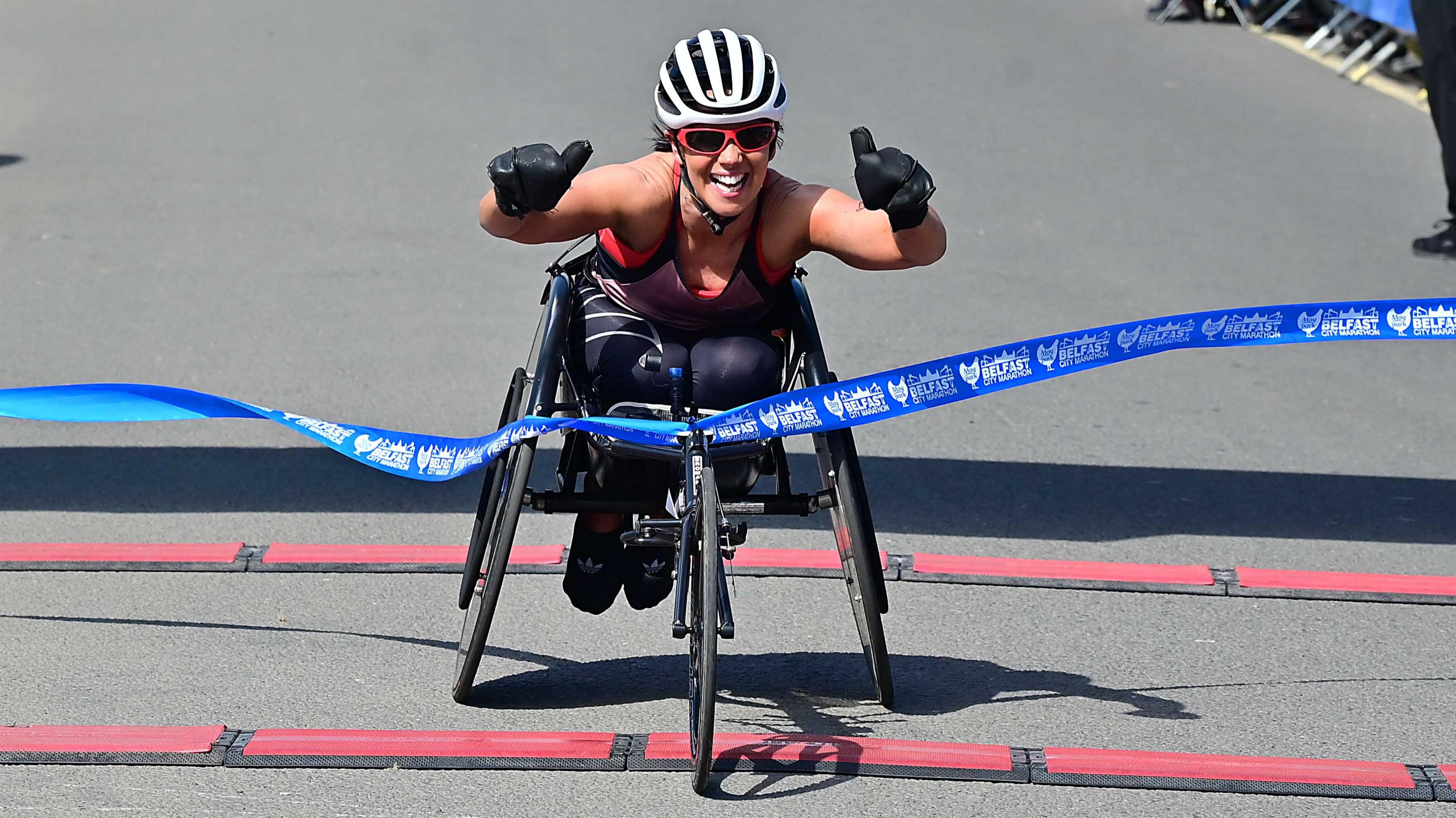 A lady in a wheelchair with a white helmet puts her thumbs up as she crosses through the blue ribbon. She is wearing red glasses and is wearing black gloves. She is wearing a vest with leggings.