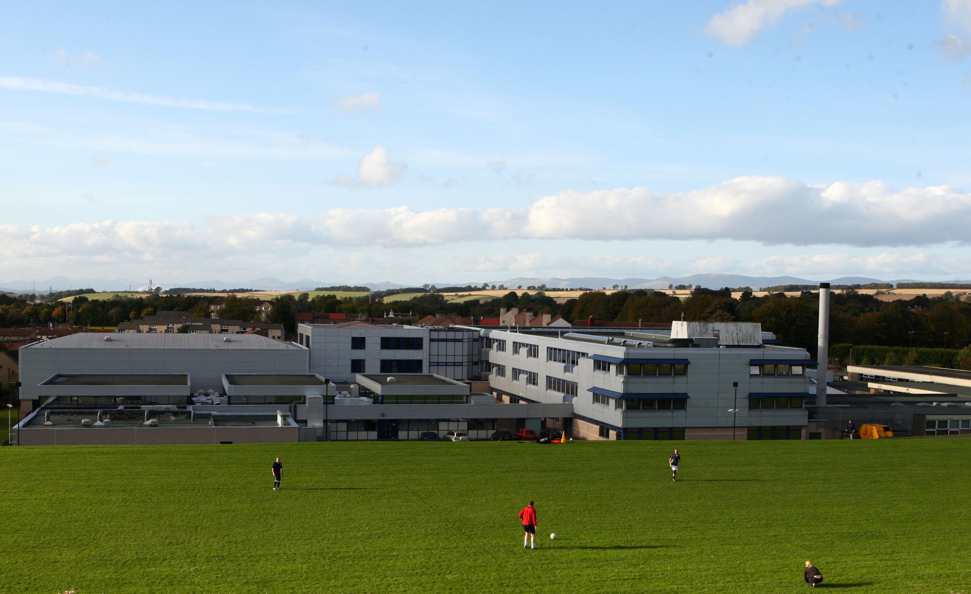 A green field with people playing football is in the foreground and in the background is a complex of two and three two storey grey buildings 