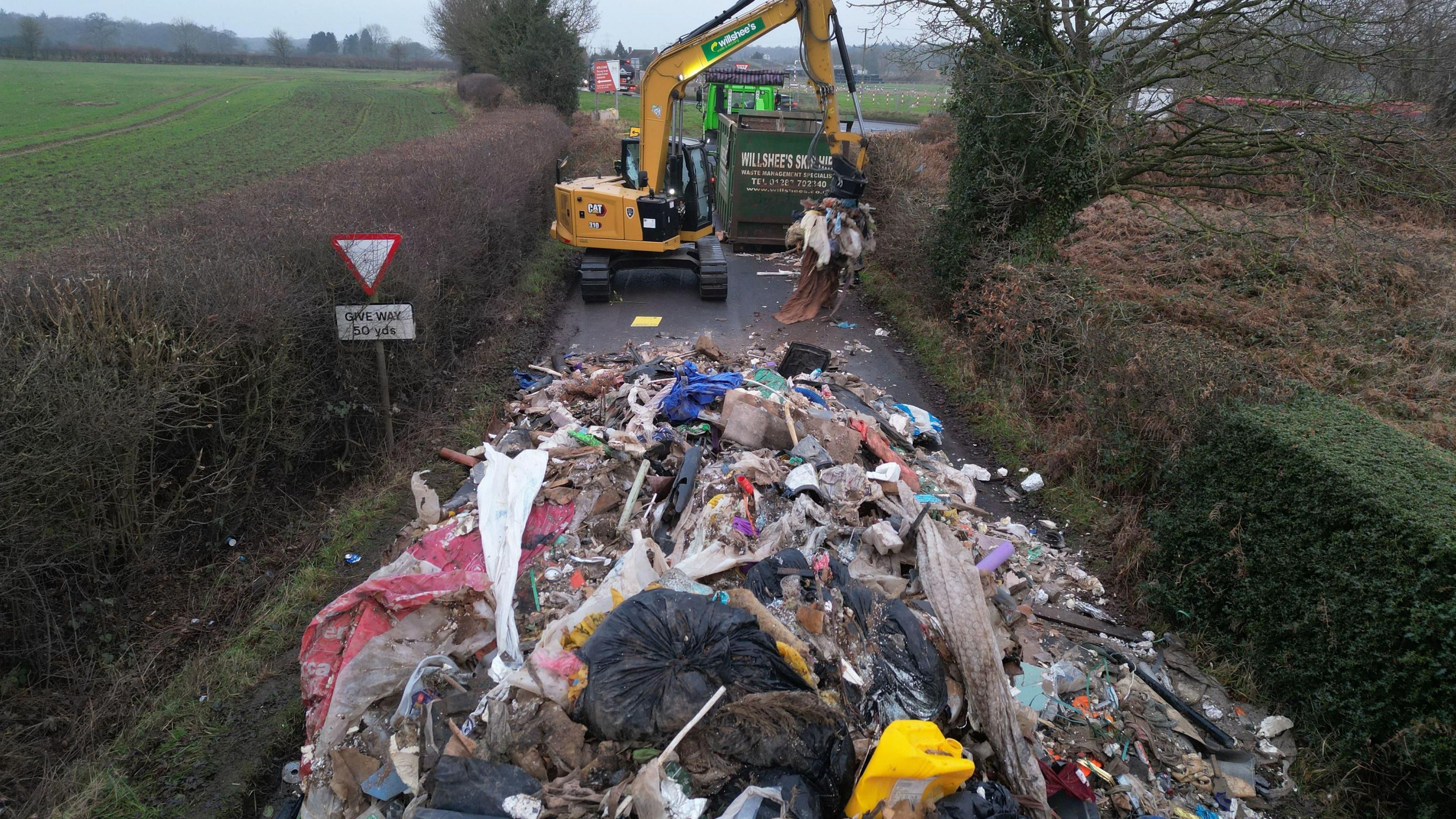 A huge pile of waste in the foreground is being handled by a yellow digger which is scooping the waste into a large green container. The pile of waste visibly blocks the road, and is close to a junction.