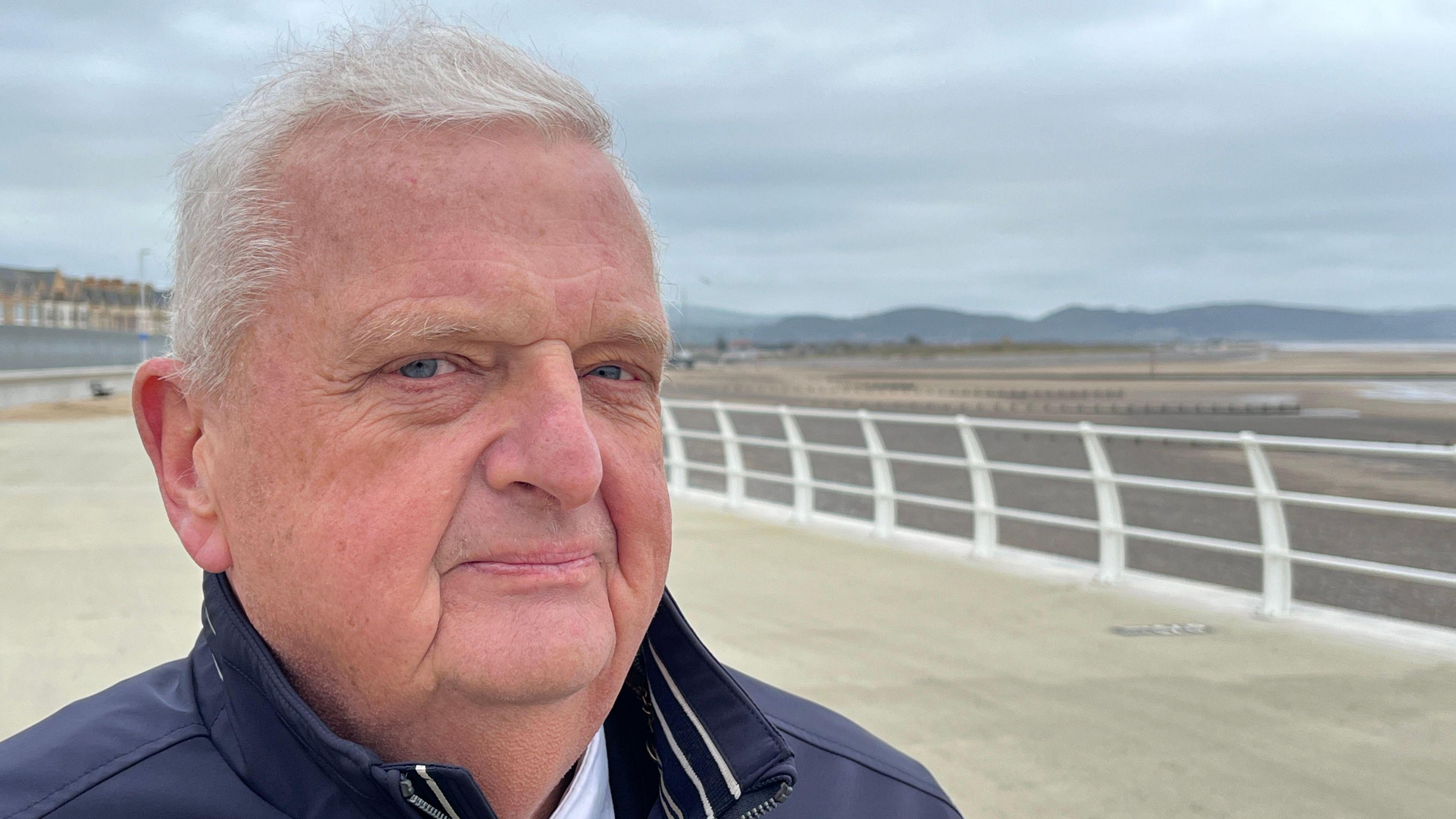 Cynghorydd Arwel Roberts o Gyngor Sir Ddinbych yn sefyll ar hyd y traeth yn Rhyl lle mae amddiffynfeydd llifogydd newydd. Mae'n gwisgo siaced las tywyll.