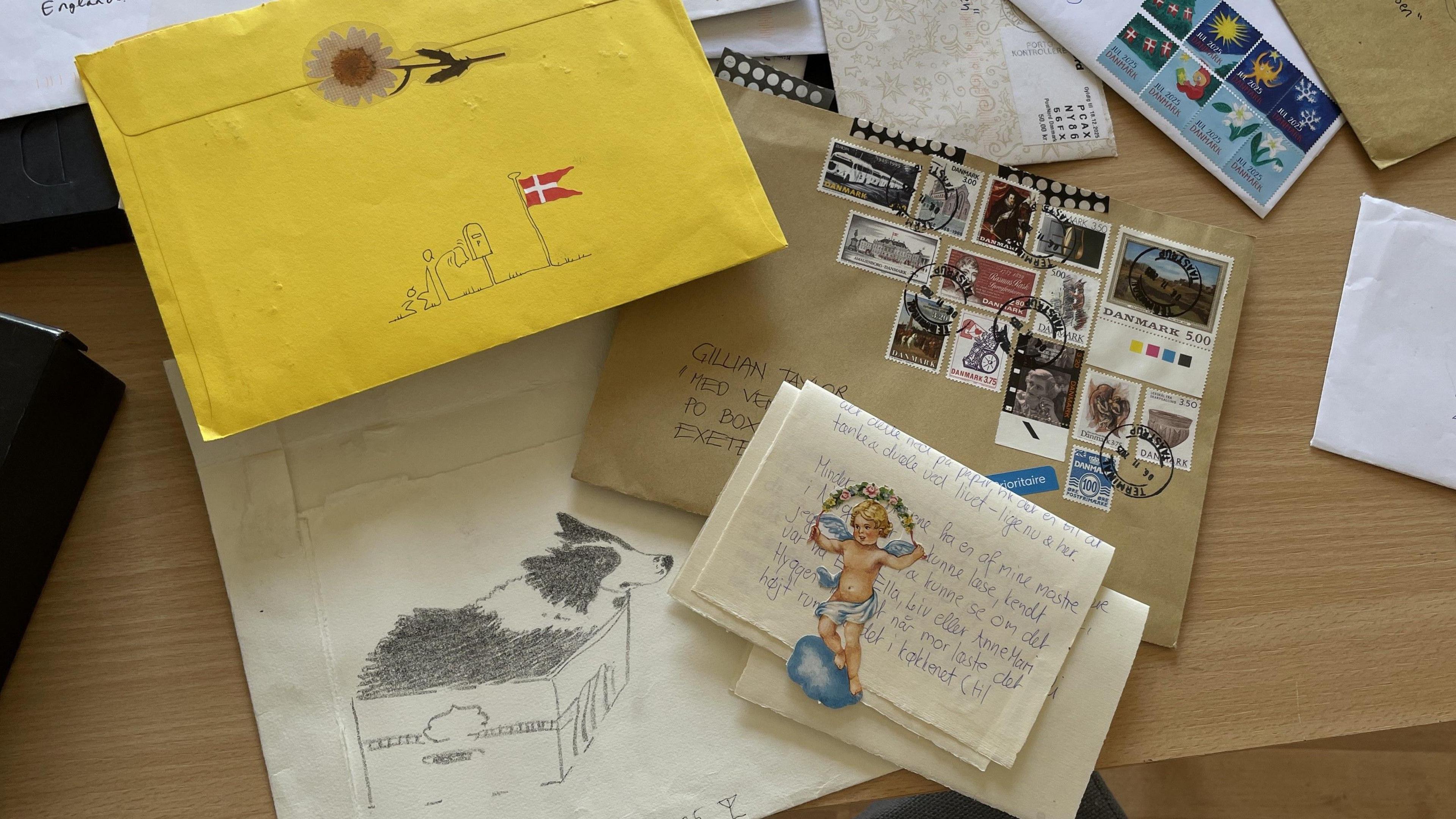 A pile of envelopes lie on the table. A yellow envelope has a line drawing of a person waving goodbye to a postbox. One envelope has a pencil sketch of a collie dog in a cardboard box. One has 14 different stamps on it. Also on the table is a folded letter with a small paper cut out of a cherub. 