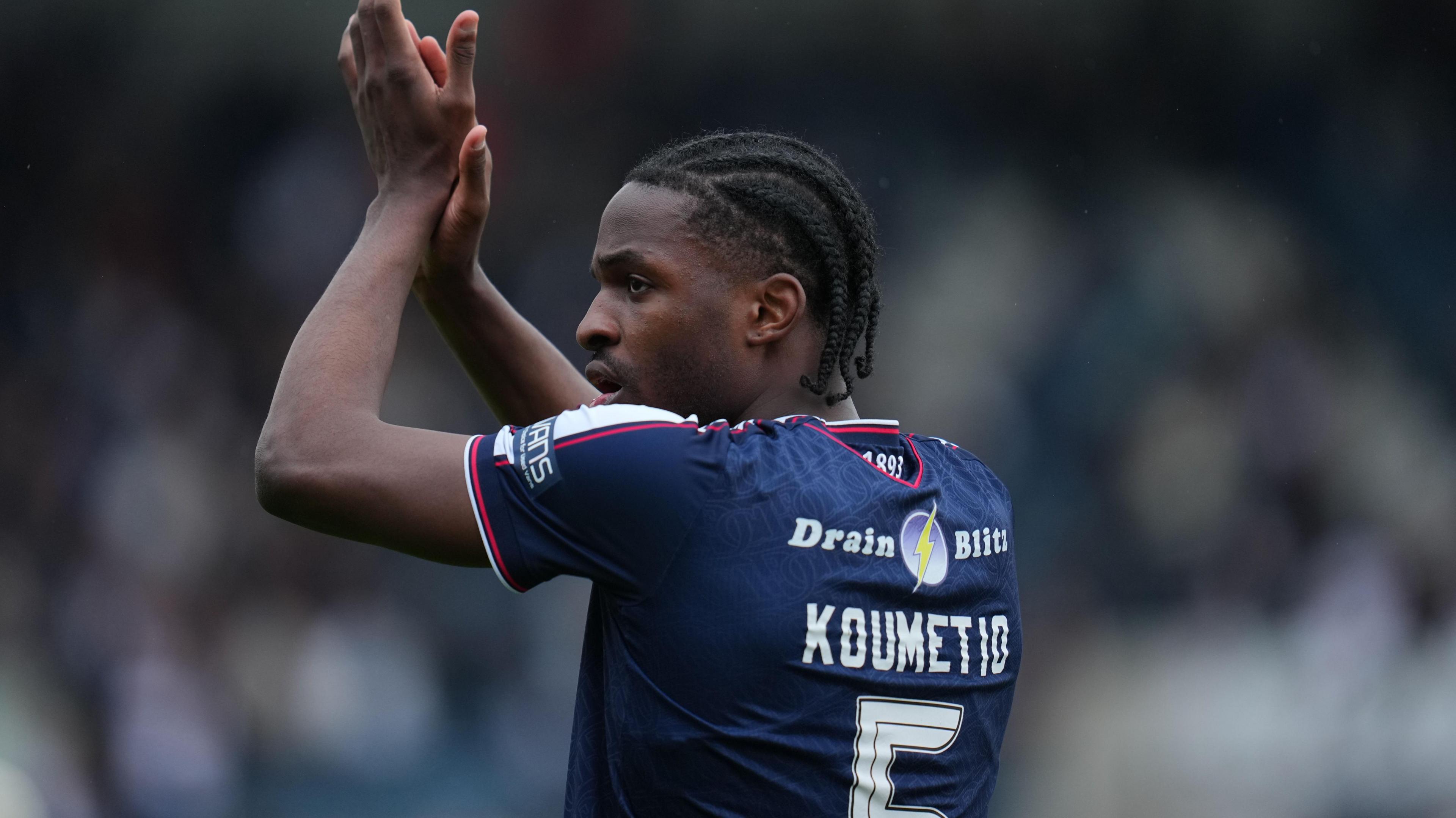 Billy Koumetio in blue Dundee shirt, facing slightly away from camera so you can see his name and number, applauds fans