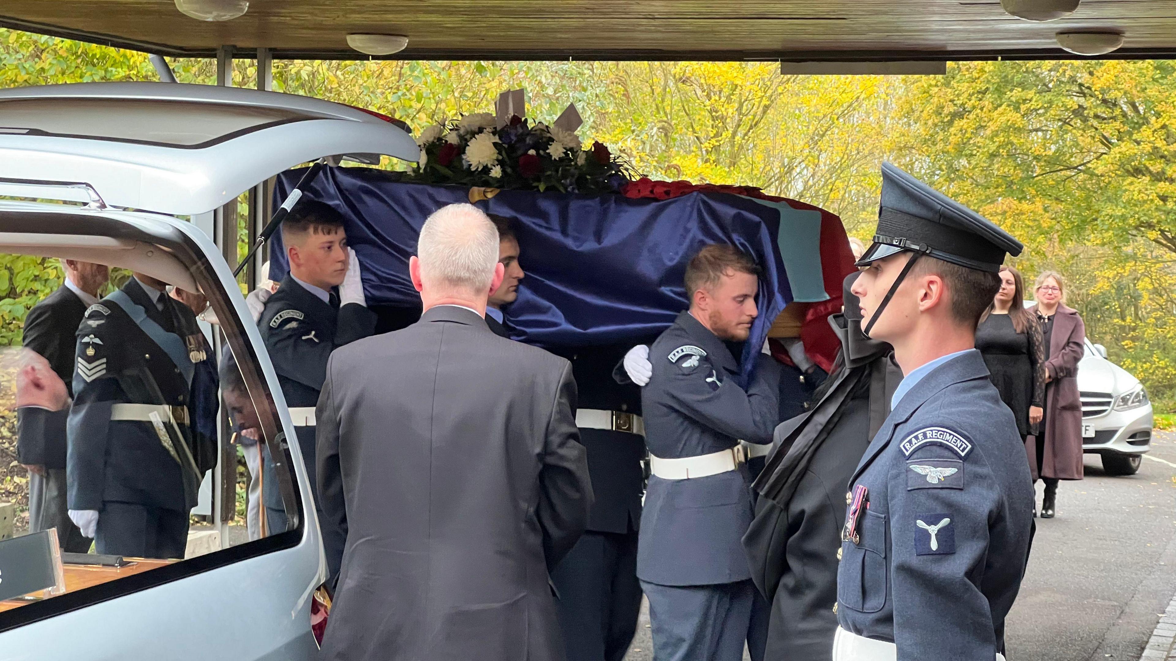 Pallbearers in blue RAF uniforms carry the flag-draped coffin into the crematorium with serious faces.