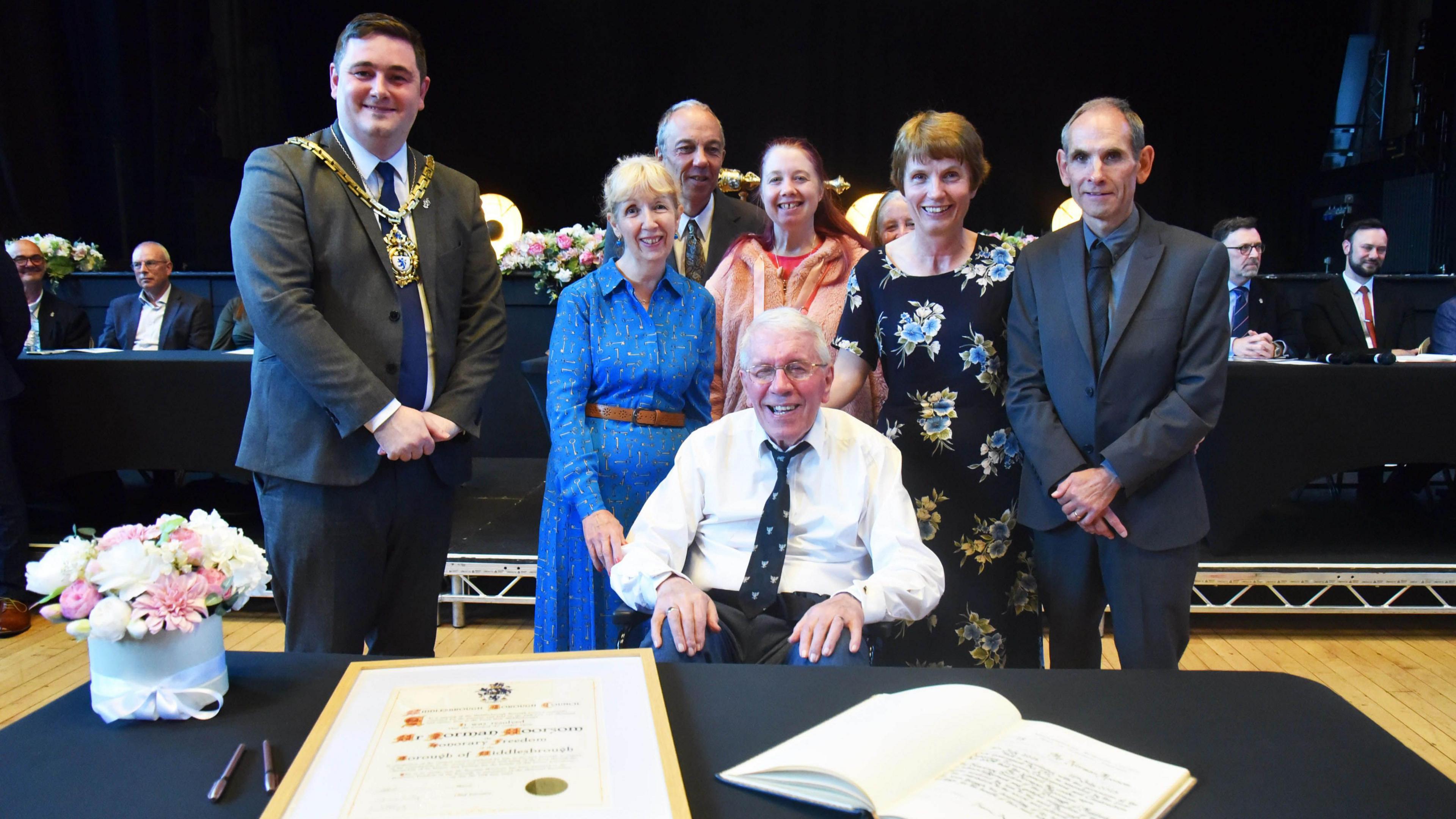 Norman Moorsom, who is seated behind a desk bearing a framed certificate, smiles into the camera. Behind, from left to right, are Middlesbrough Mayor Chris Cooke, Mr Moorsom's friend Linda Ford, his son Richard Moorsom, granddaughter Laura Moorsom, daughter Hilary Webb and son-in-law Martin Webb.