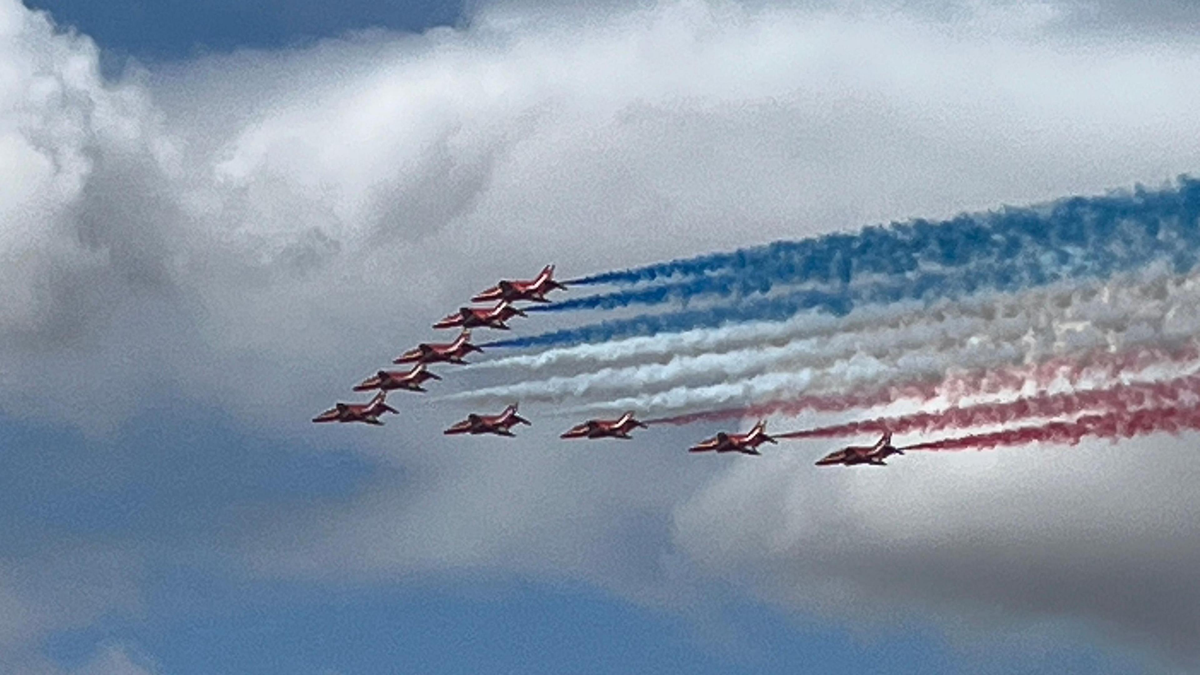 A fleet of red planes organised in a pointing pattern with blue, white and red fog coming out of their rears. They are flying in the sky with clouds in the background.