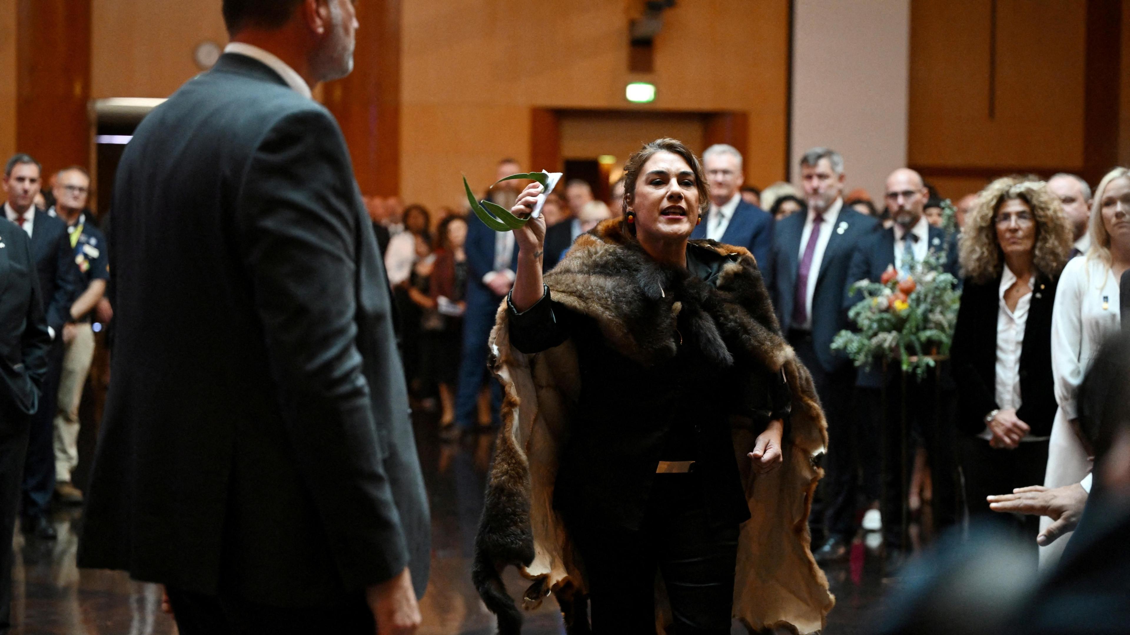 A woman wearing a possum skin cloak gesticulates as she stands amongst a crowd of people