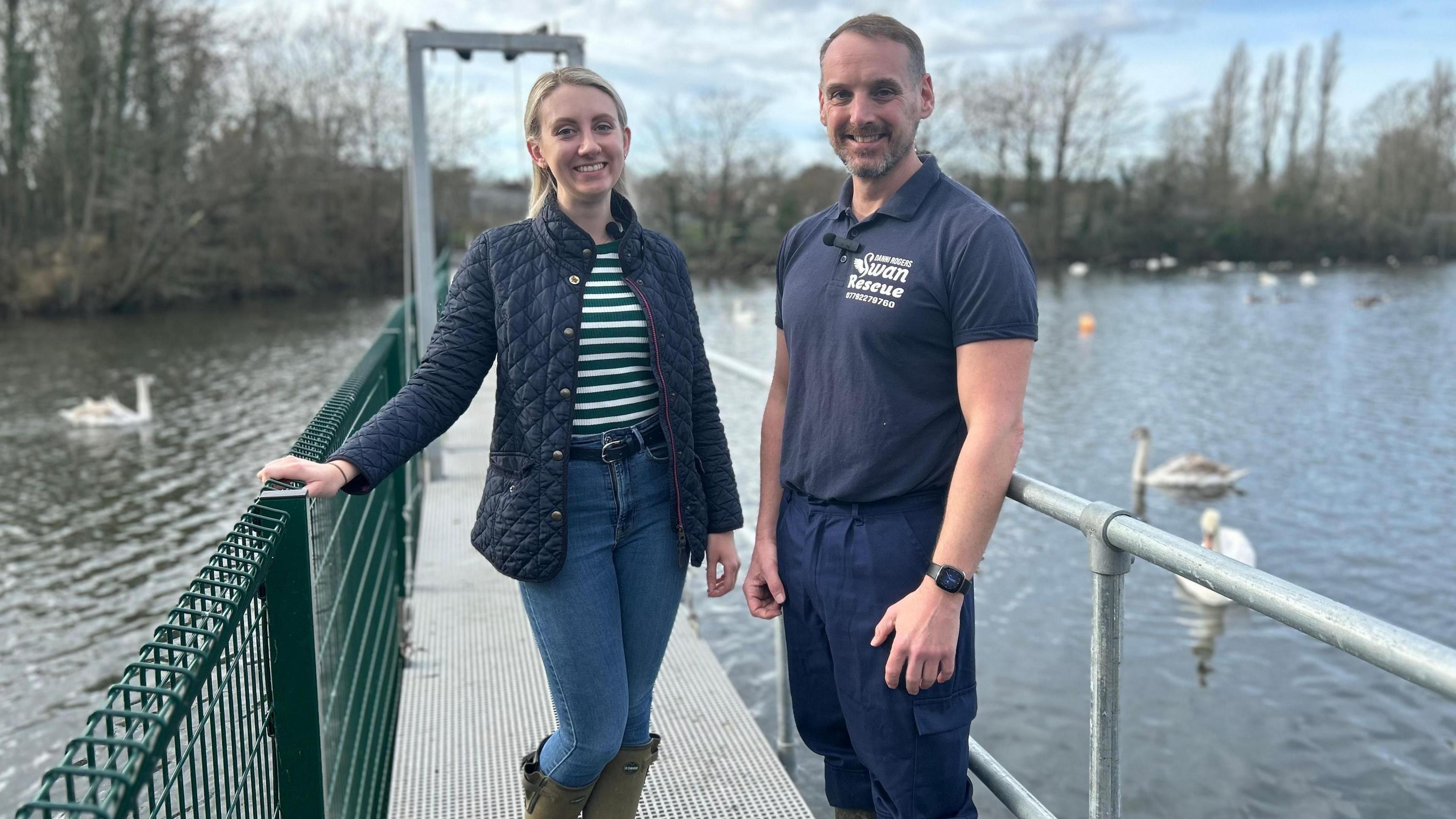 Ellie Vesey-Thompson and Danni Rogers standing on a pier at Shepperton Swan Sanctuary, with three swans swimming in the background.