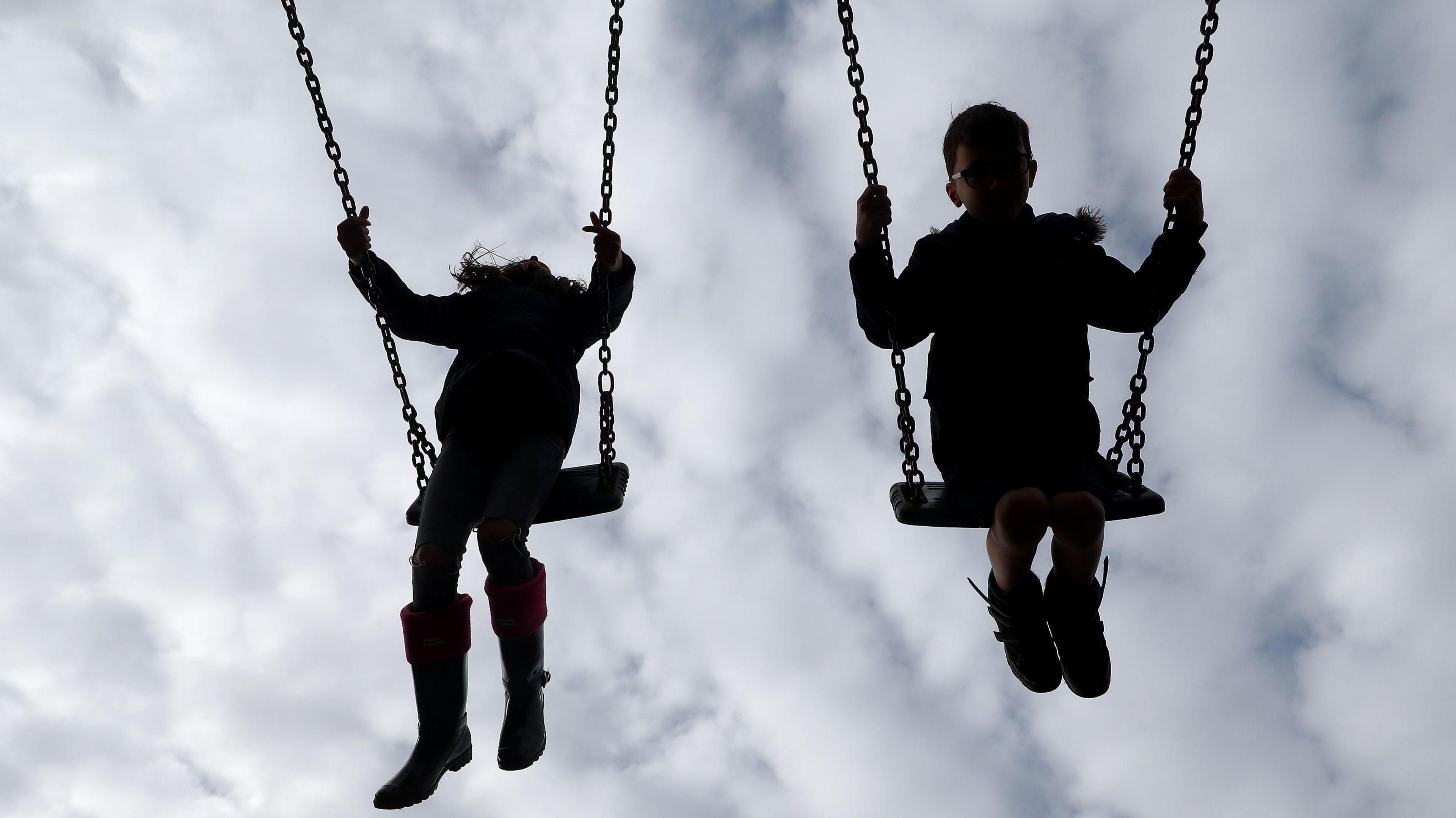 Children on swings. The two children are swinging into the air side by side. Their features are not visible and they appear silhouette-like. It appears to be a young girl on the left and a young boy on the right. The sky behind them is overcast.