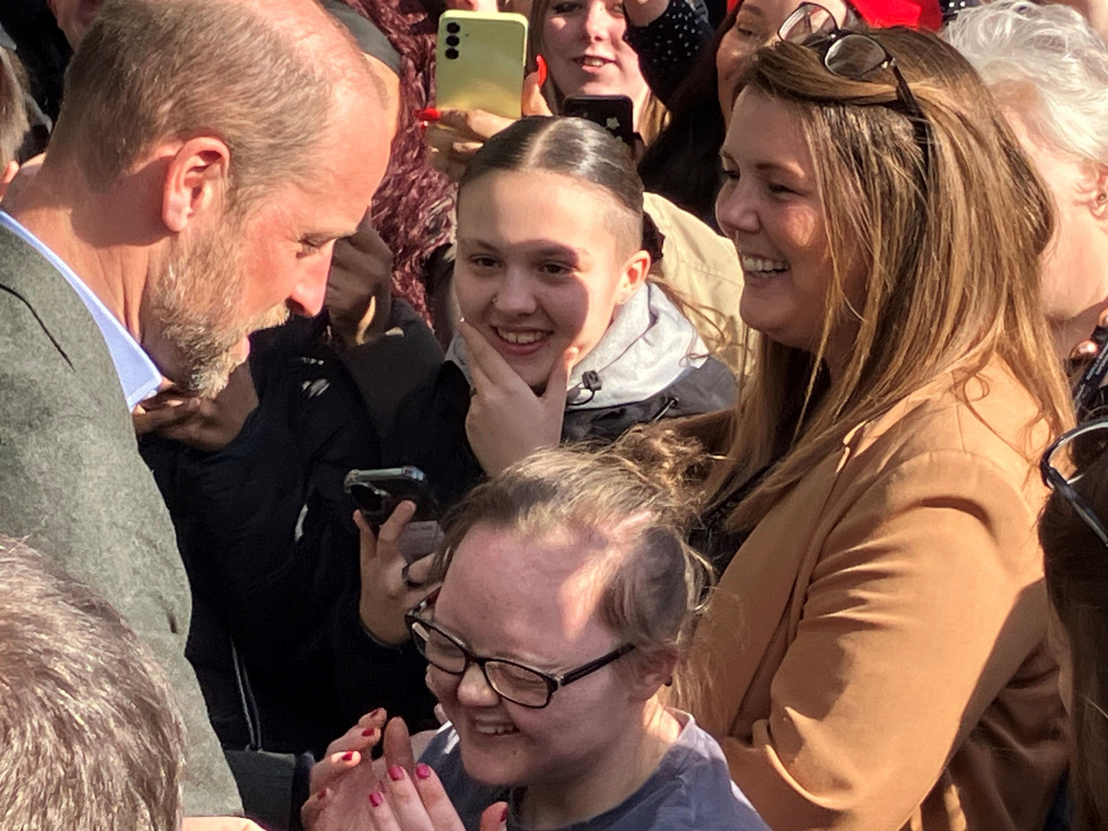 In the aftermath of the hug - a shocked Natasha Gorry holds her hands up to her face and laughs. Prince William, who is considerably taller appears to be talking to her as he smiles. Ms Gorry is wearing black rimmed glasses and her light brown hair is tied back. She is standing at the front of a crowd of people who are all smiling.