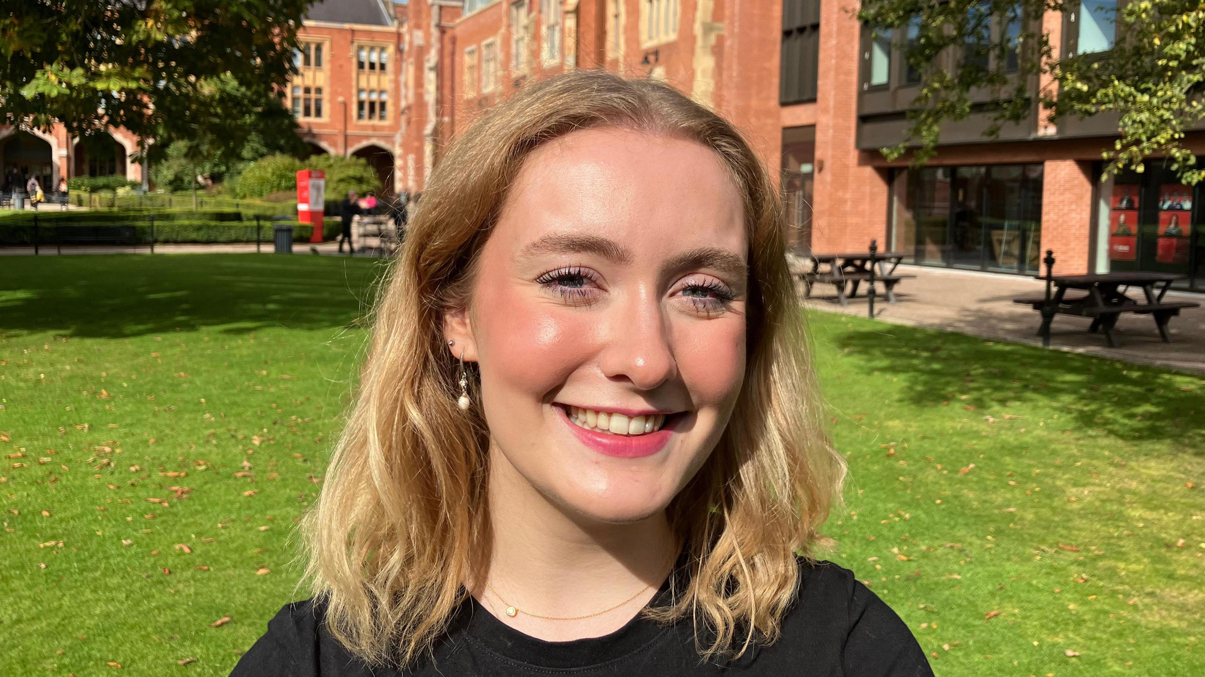 Anna is standing in front of red brick, with benches in the background. It is very sunny. 

She has blond hair, pearl earrings and a fine necklace. She has blue eyes and is wearing mascara. 

She is smiling at the camera and looks like she is wearing red lipstick. 