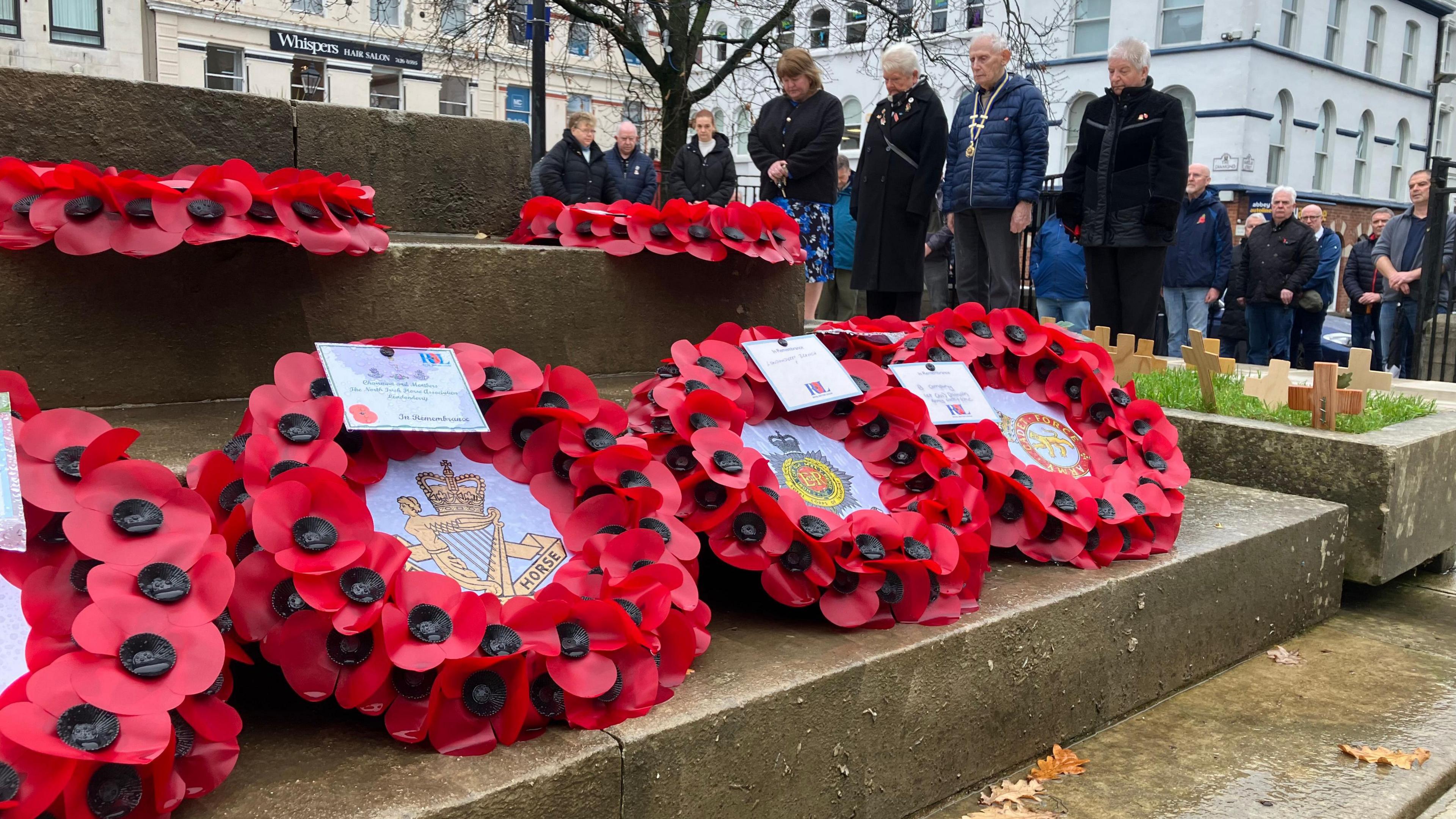 People are standing solemnly- outside in coats and formal attire at an armistice ceremony. Poppy wreaths can be seen on the steps up to the memorial. Buildings can be seen in the background. 