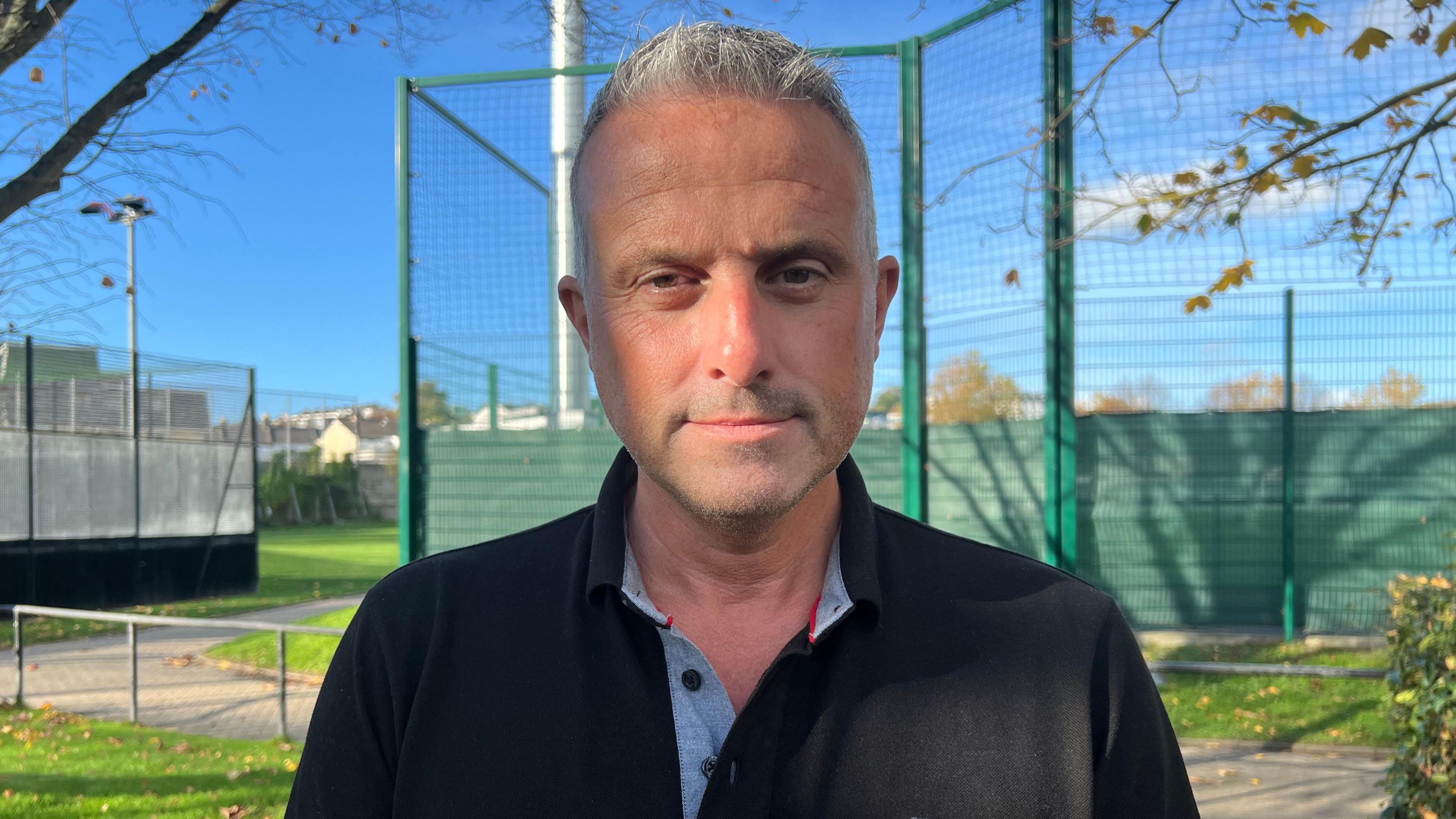 Carl Walker, a man with short grey hair and stubble. He is wearing a black polo shirt with a grey inner collar. He is staring at the camera. Behind him is a large green net and floodlights, while grass is visible to the left of the image.