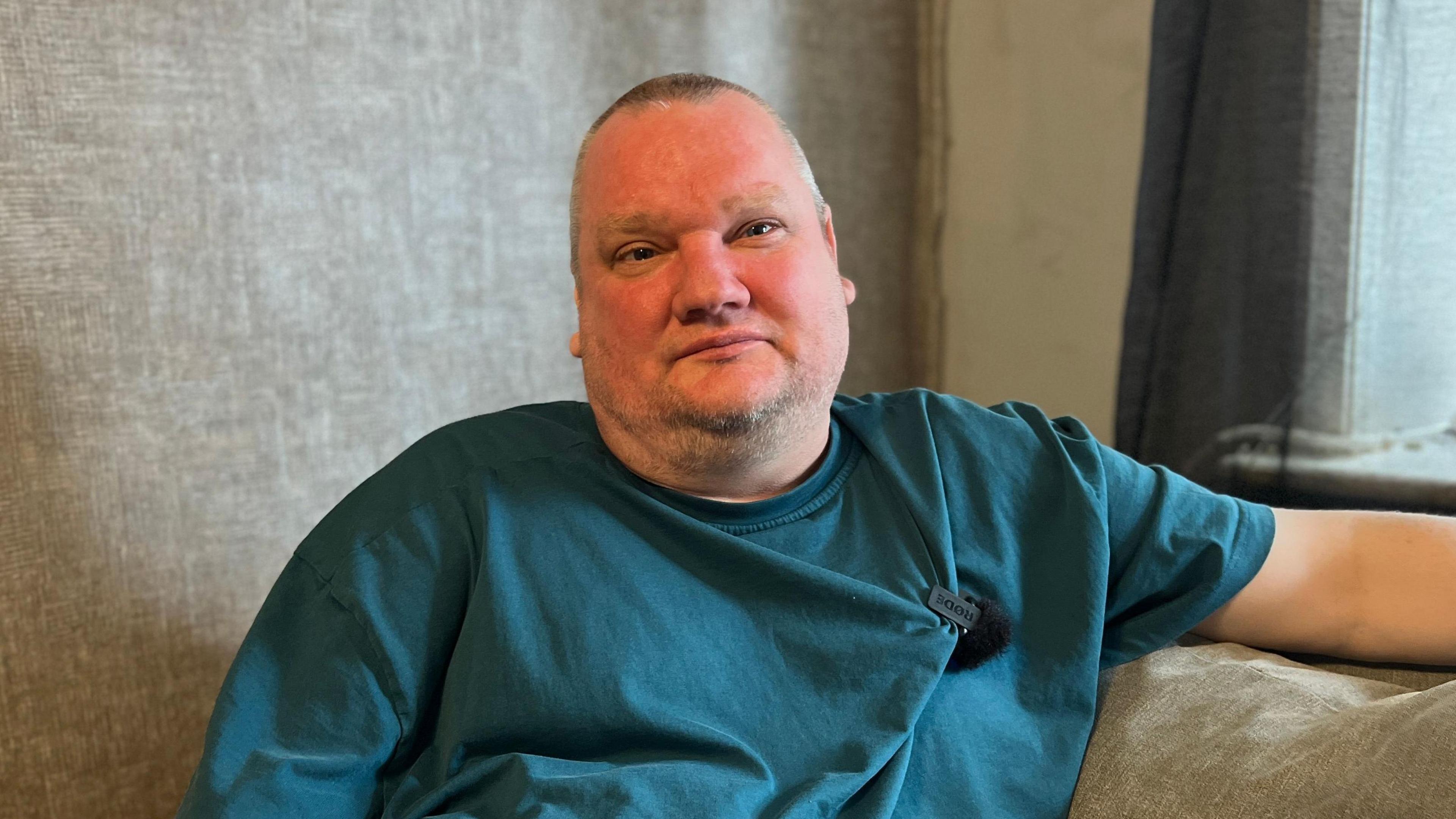 Shaun Pinkney, wearing a blue t-shirt, sits on the sofa in his living room