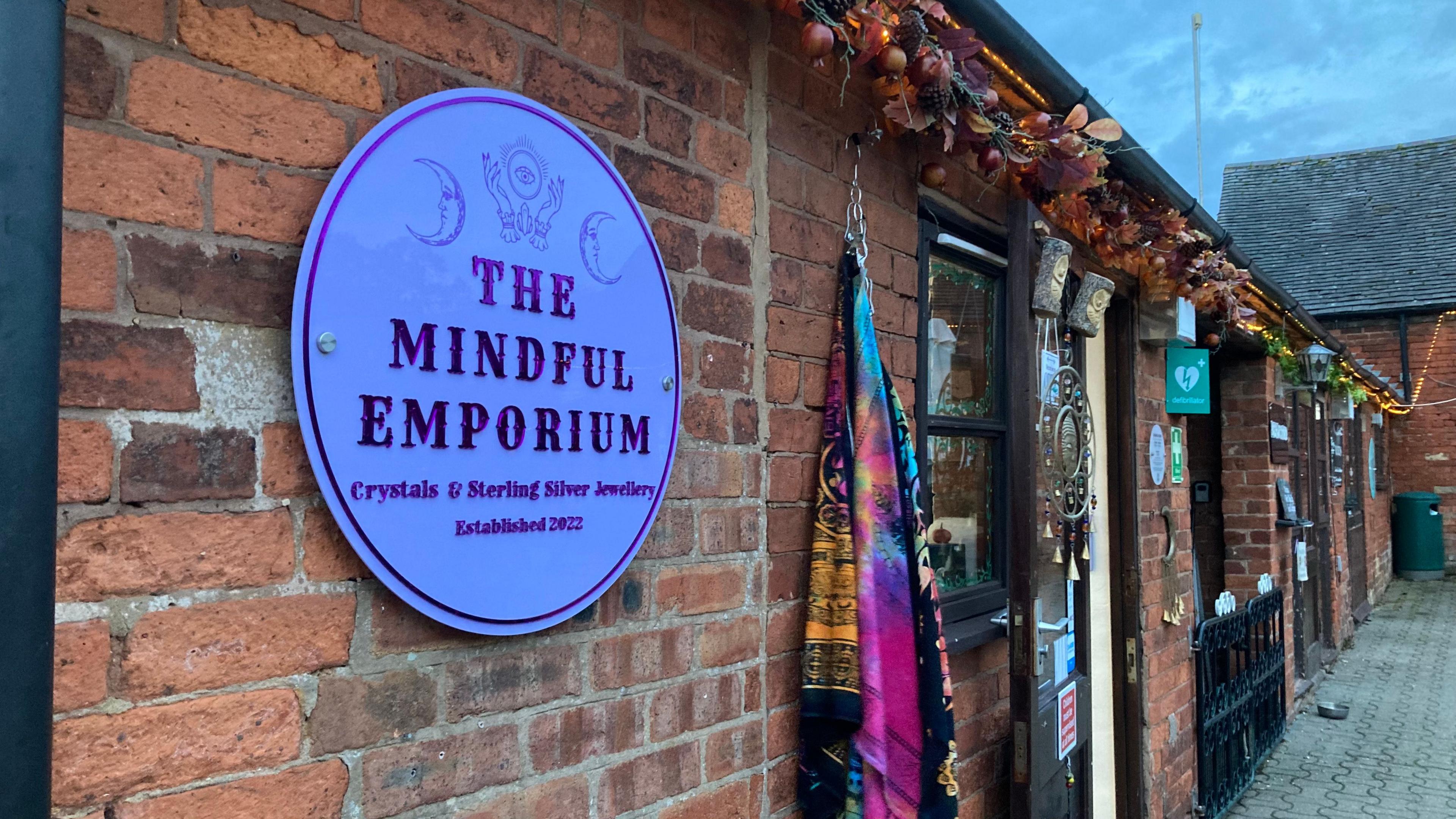 A circular mauve sign is attached to a a building with red brick wall, the sign has Mindful Emporium written on it. There is a purple and black scarf attached next to an open door.