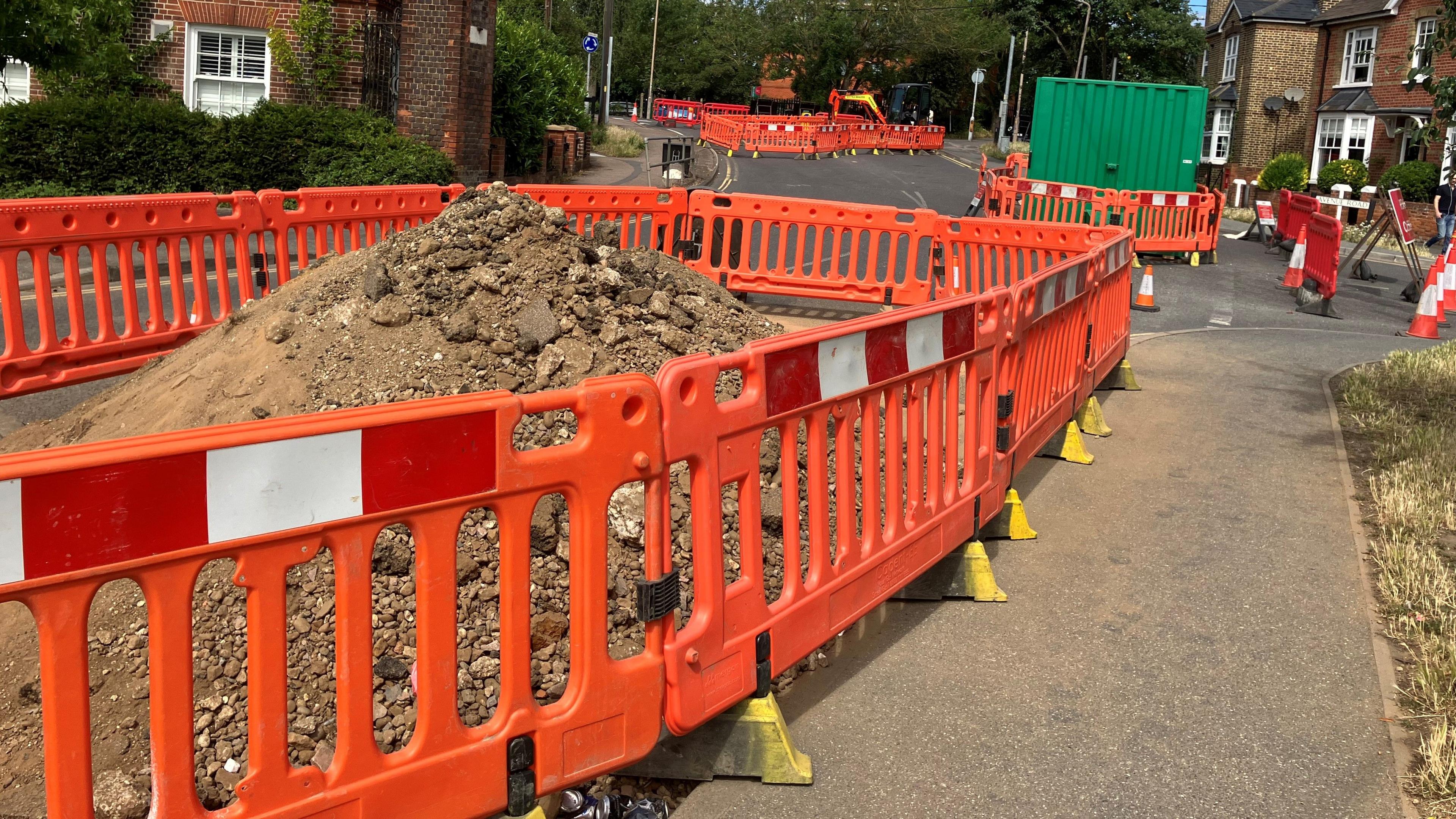 A big mound of dirt surrounded by orange fencing, more fencing and cones are visible further up the road
