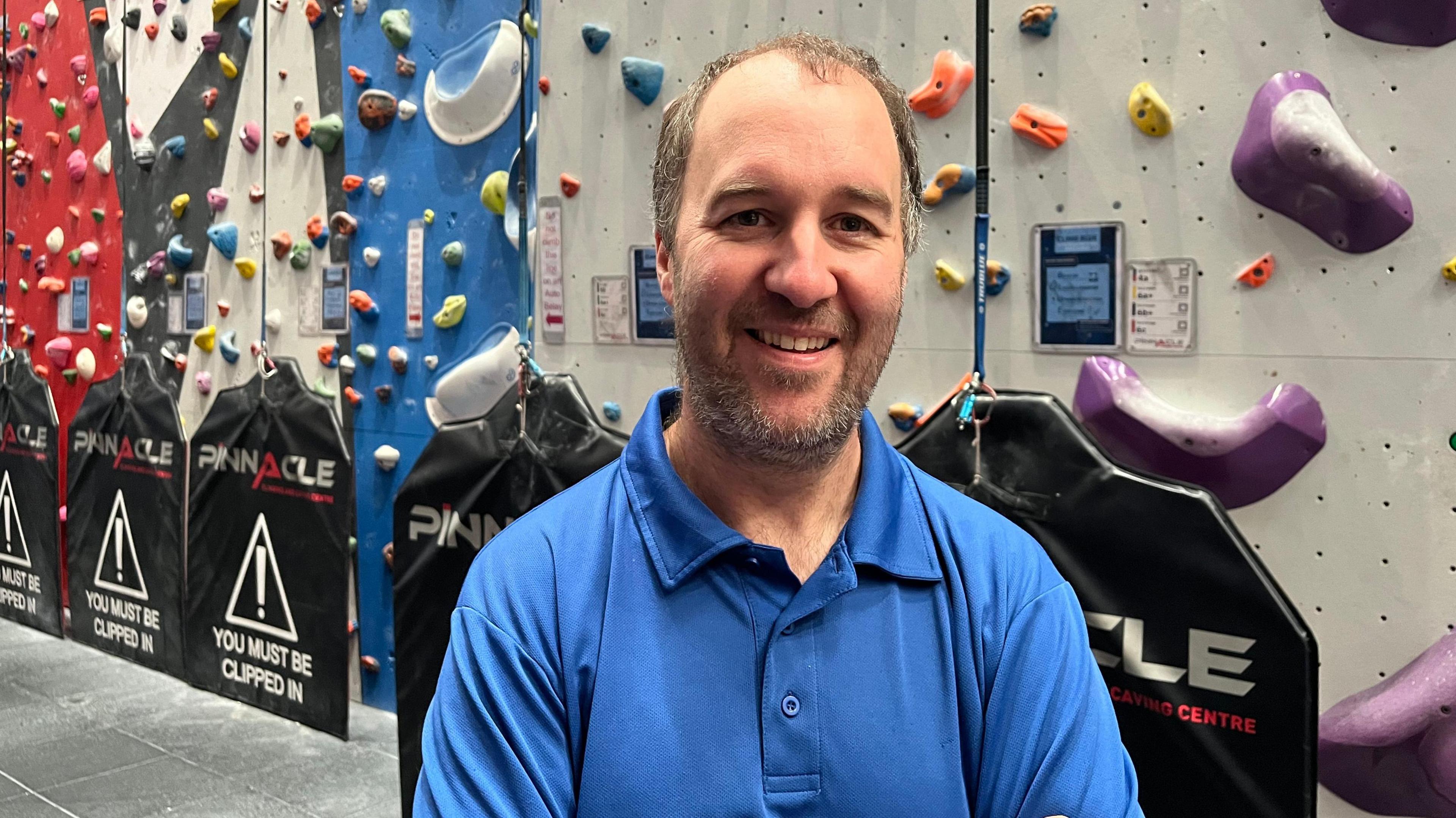 A man with a short beard wearing a blue polo shirt stands in front of climbing equipment. 