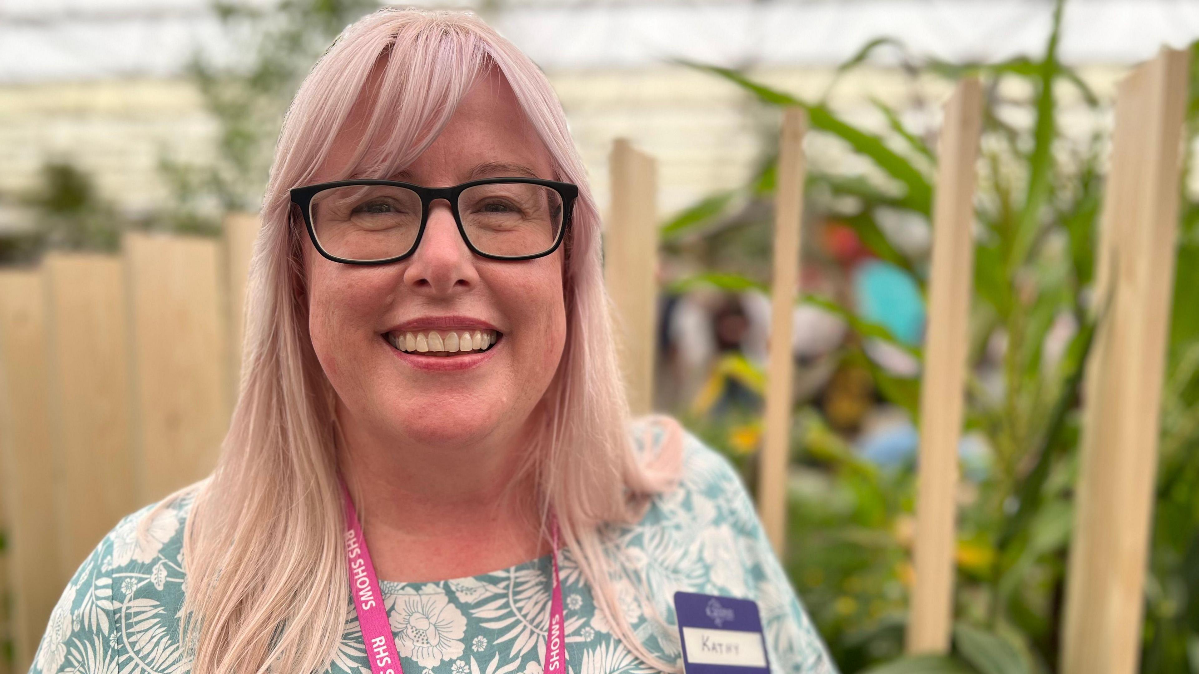 Kathy Grube has blonde and pink hair. She is standing in front of an RHS Chelsea Flower Show exhibition. She is wearing a blue floral outfit, a pair of glasses and is looking at the camera and smiling.