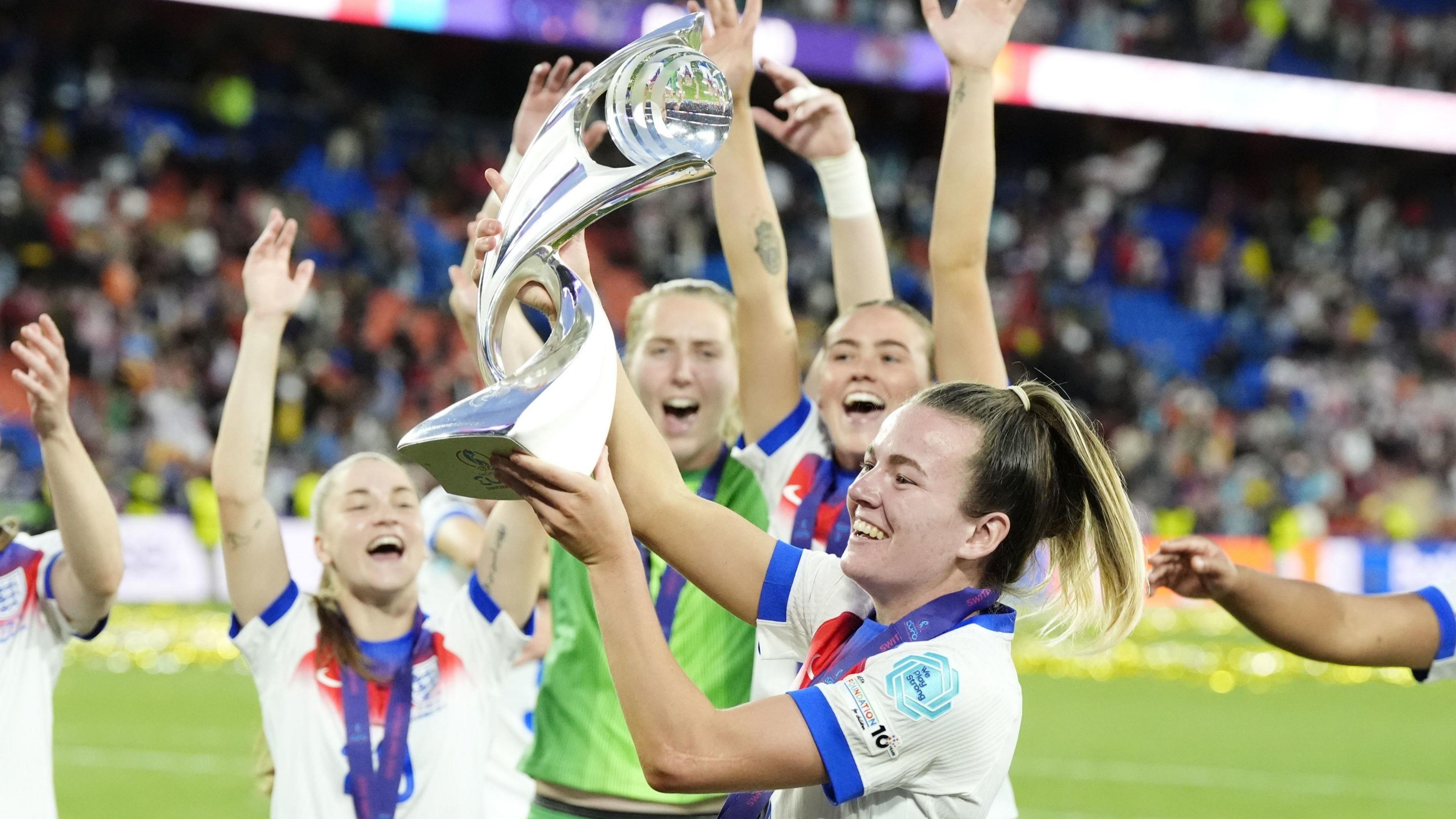 A female footballer lifts a trophy in the air as her team-mates cheer behind her.