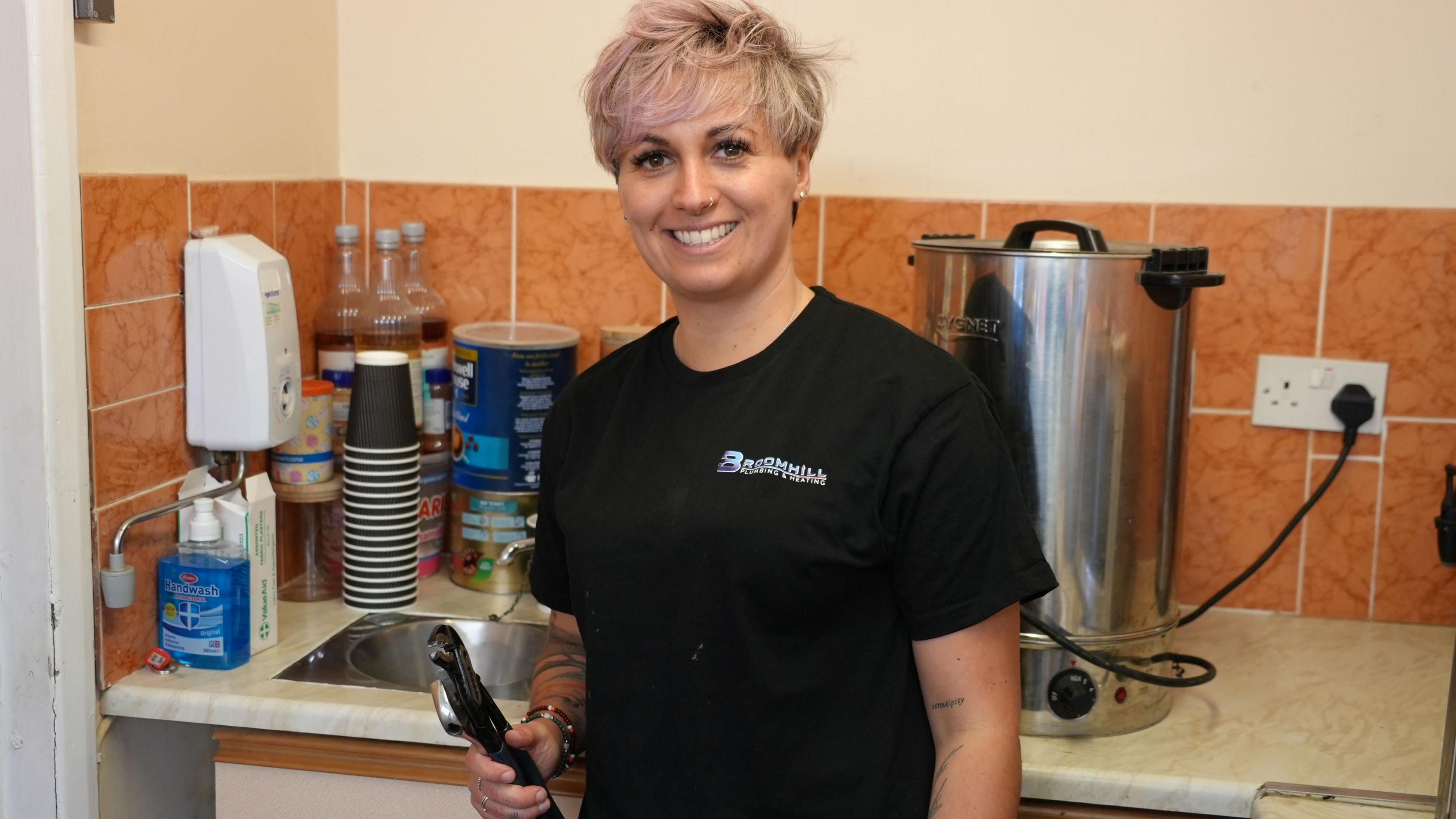 Woman is smiling and has pink hair. She is in a kitchen with a tea urn and paper cups and a sink behind her. She is wearing a black T-shirt and holds a large spanner.