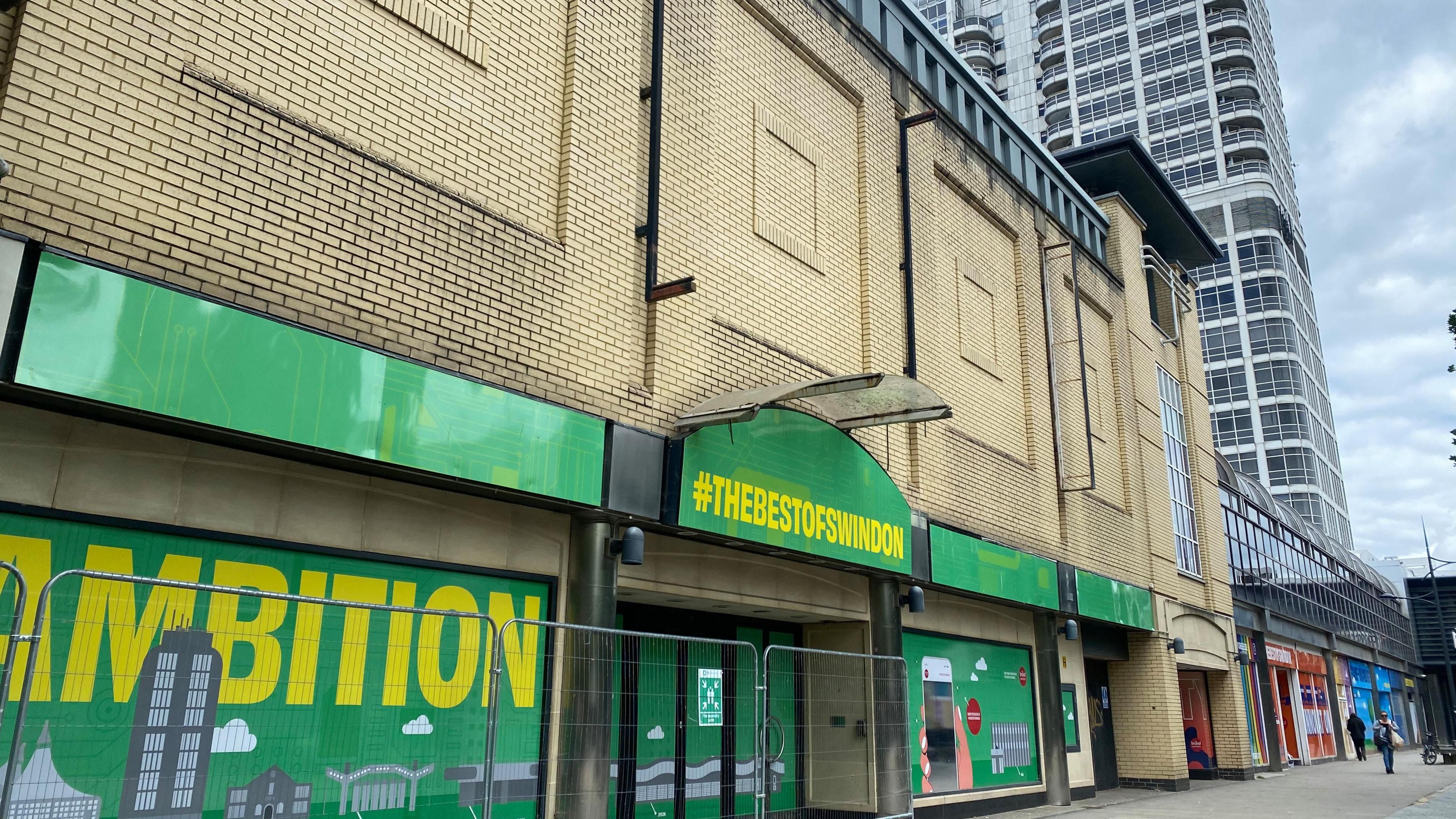 The centre of Swindon with the David Murray John tower block in the background and a new build - but empty - shop with railings in front of it. It has a green sign saying #thebestofswindon