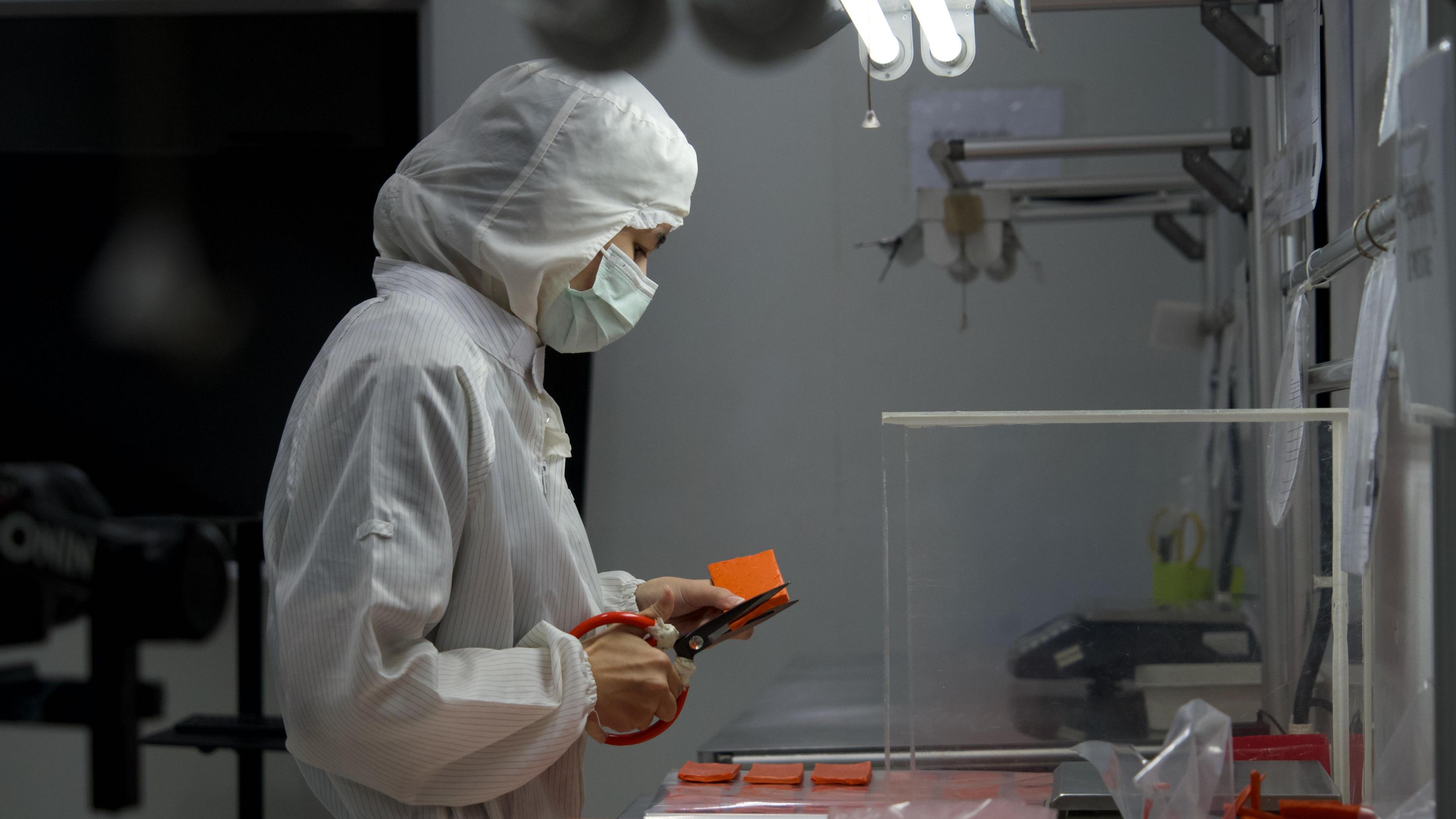 A woman in overalls with her head covered and wearing a mask is cutting rubber in a factory in Thailand.