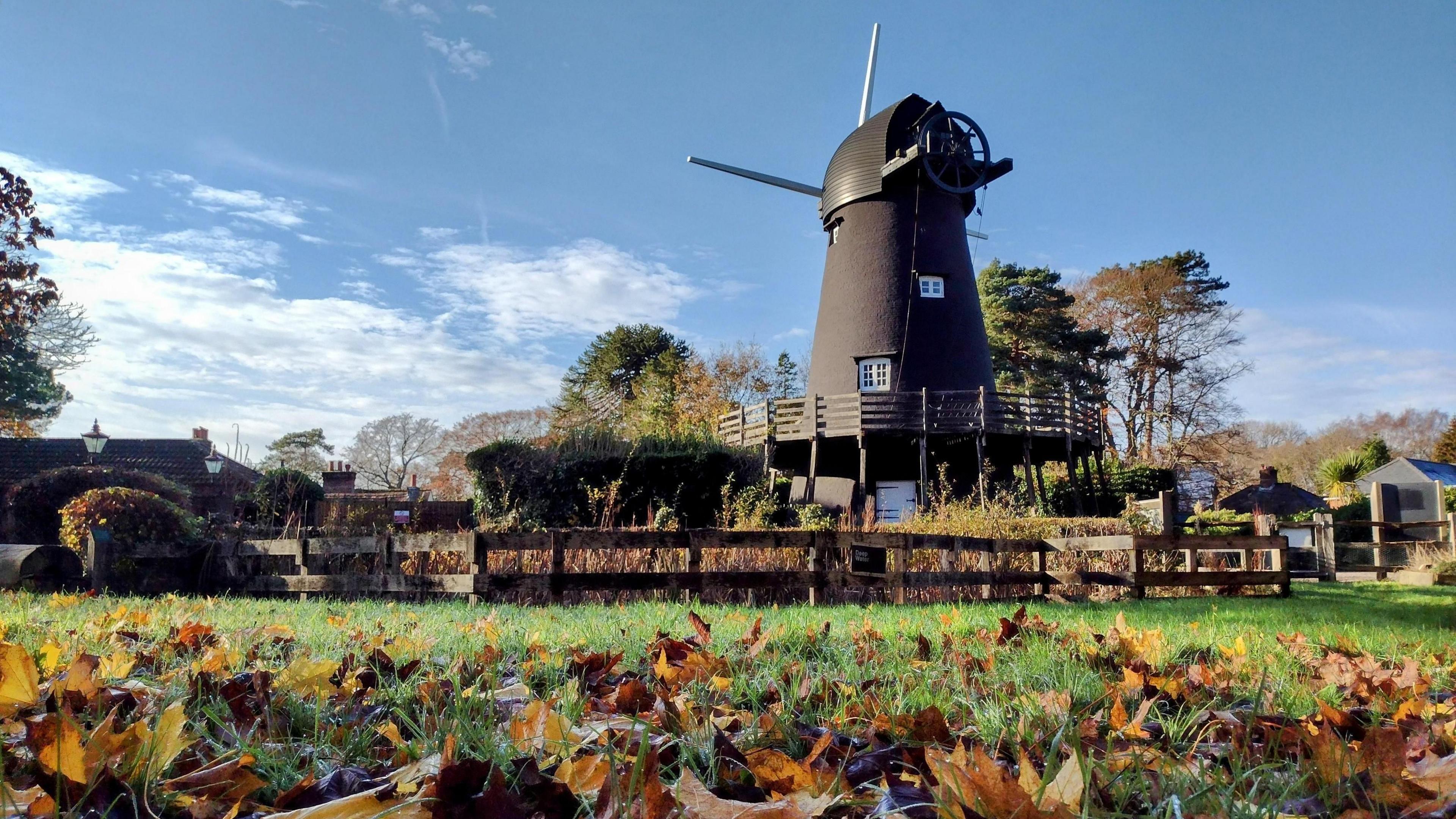 A windmill is the main focus of the picture with a green field carpeted with leaves in front of it and a blue sky with wispy clouds behind it. There are several windows inset in the windmill and you can make out two of its sails.