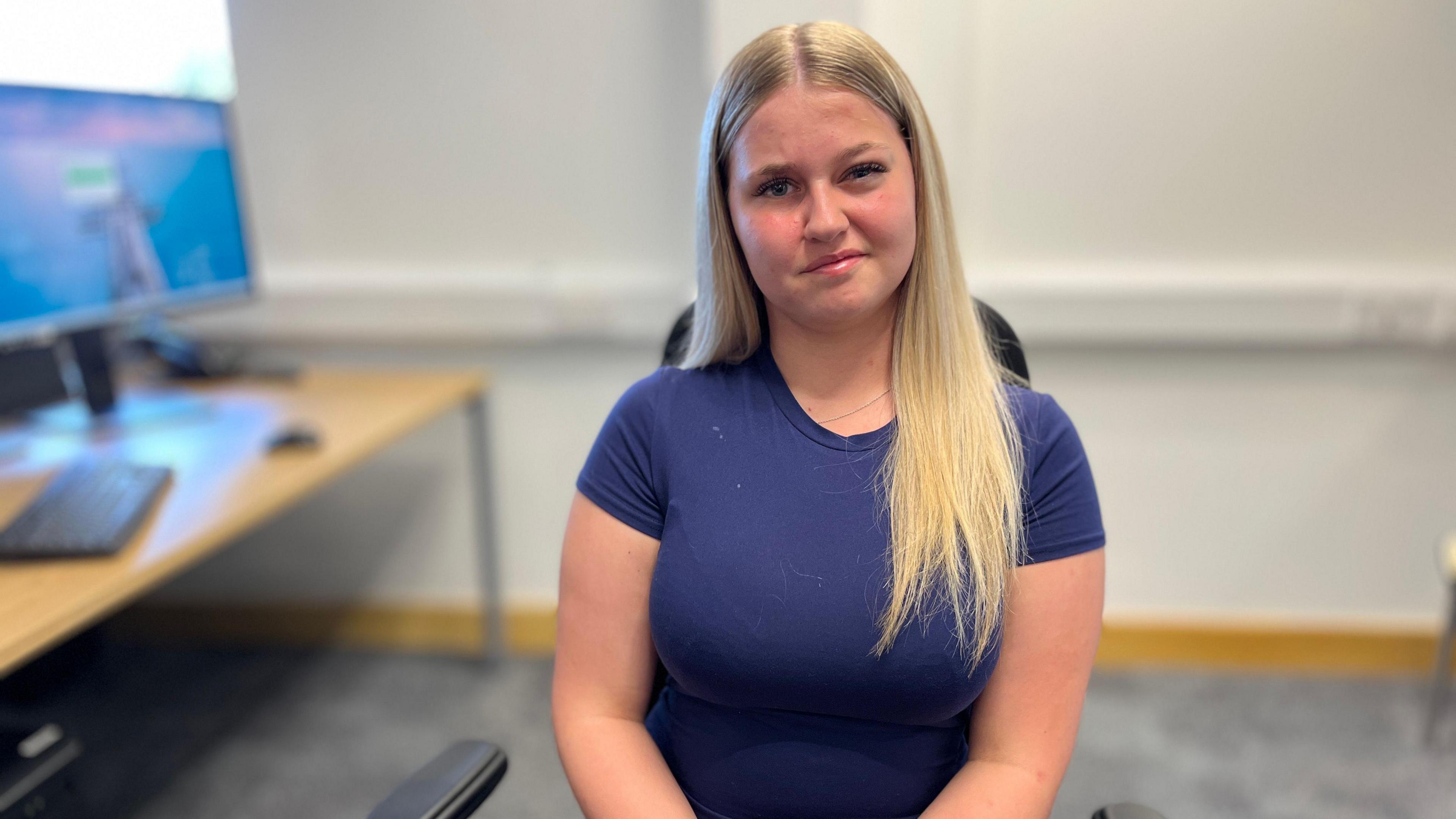 A teenage girl with long blonde hair is sitting in an office wearing a navy blue T-shirt. There is a computer on a desk to her right.